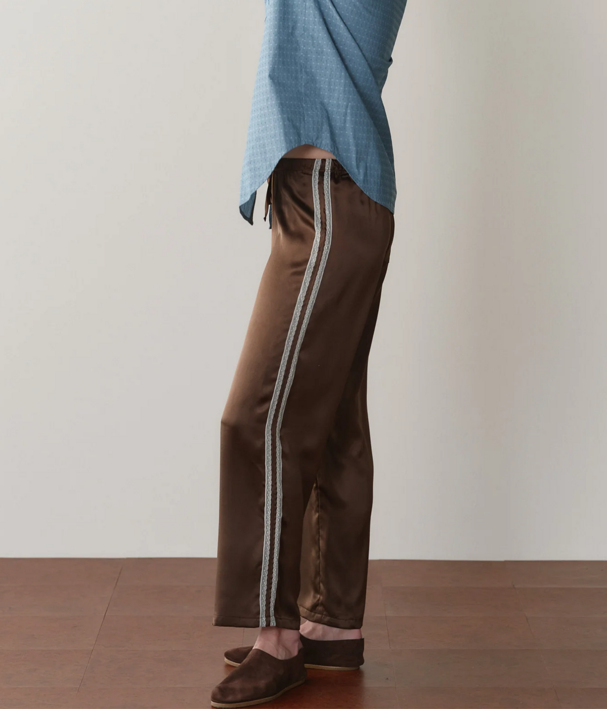 Three-quarter side view of a model wearing the DONNI. The Silk Lace Stripe Crop Pant in Espresso, showing the lace trim running to the hem, with a blue printed shirt and brown suede slippers