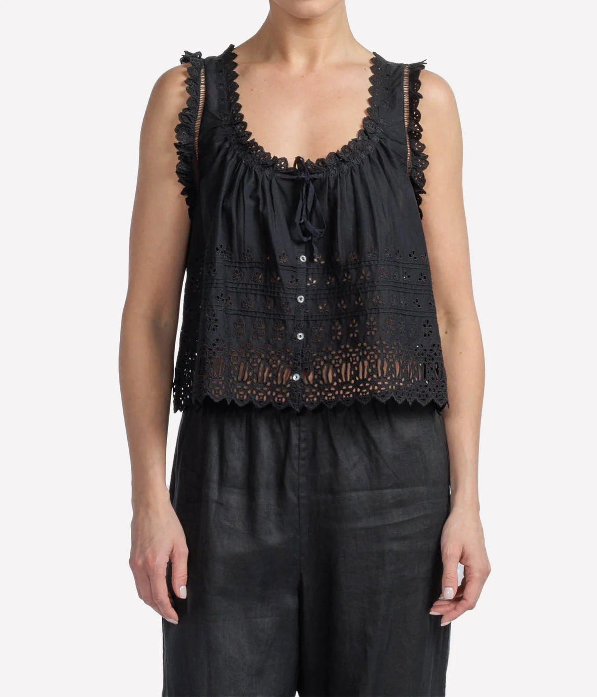 A woman wearing a black sleeveless eyelet lace top with ruffled trim and a drawstring neckline, paired with black pants, against a light gray studio background.