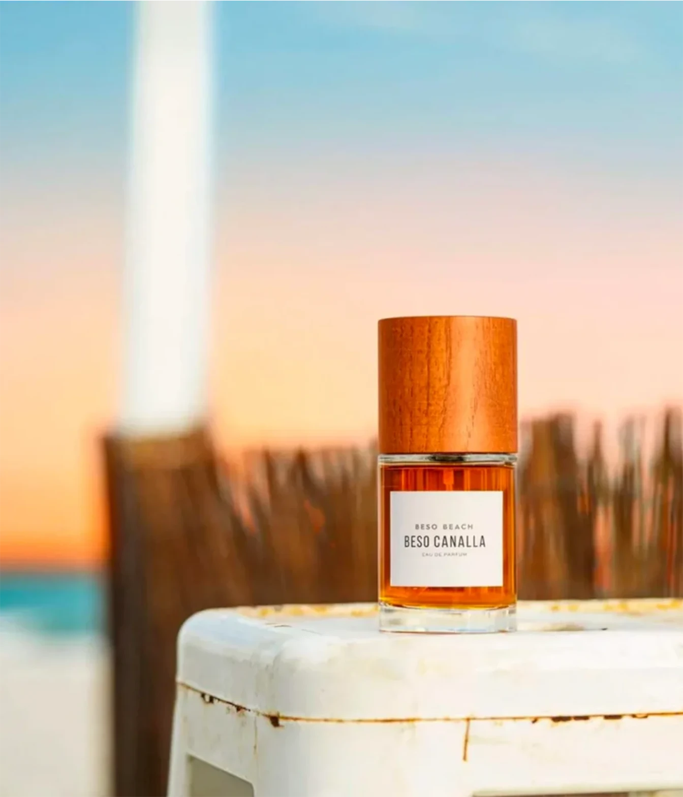 A bottle of Bego Canalla 'Nido Beach' perfume with a wooden cap and amber liquid, displayed on a white stool with a blurred beach sunset background.