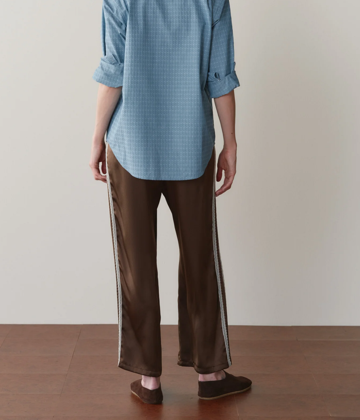 Back view of a model wearing the DONNI. The Silk Lace Stripe Crop Pant in Espresso with a blue printed shirt with rolled sleeves and brown suede flats, showing the relaxed straight fit