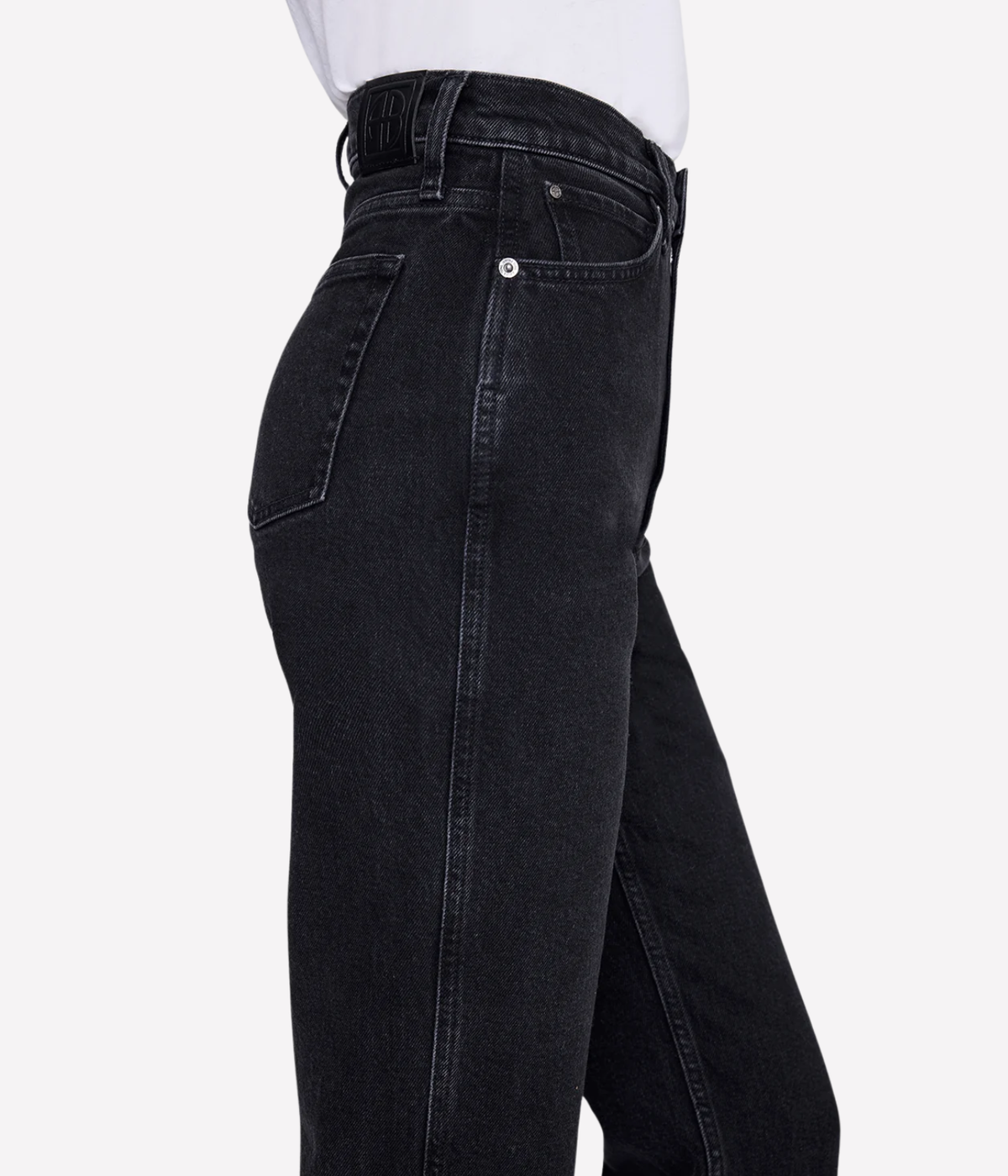 Close-up side view of a person wearing black denim jeans with a white top visible at the waist, against a light gray studio background.