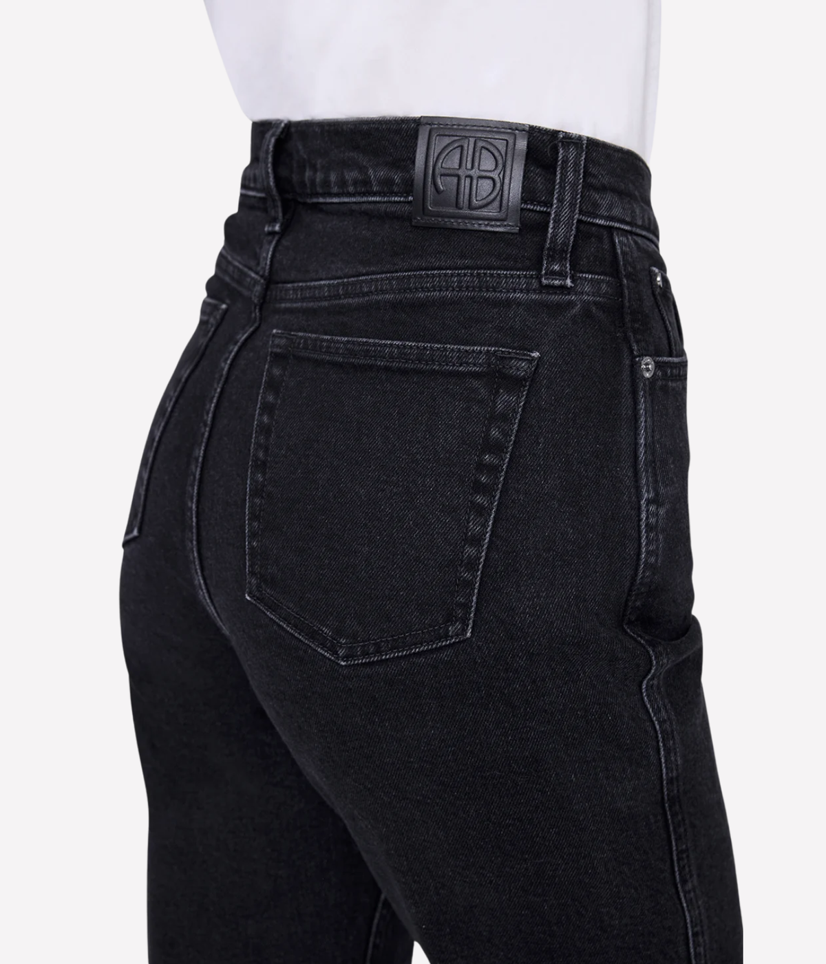 Close-up rear view of a person wearing black denim jeans with a white shirt at the waist, showing the back pocket, stitching, and a black leather brand patch on the waistband.