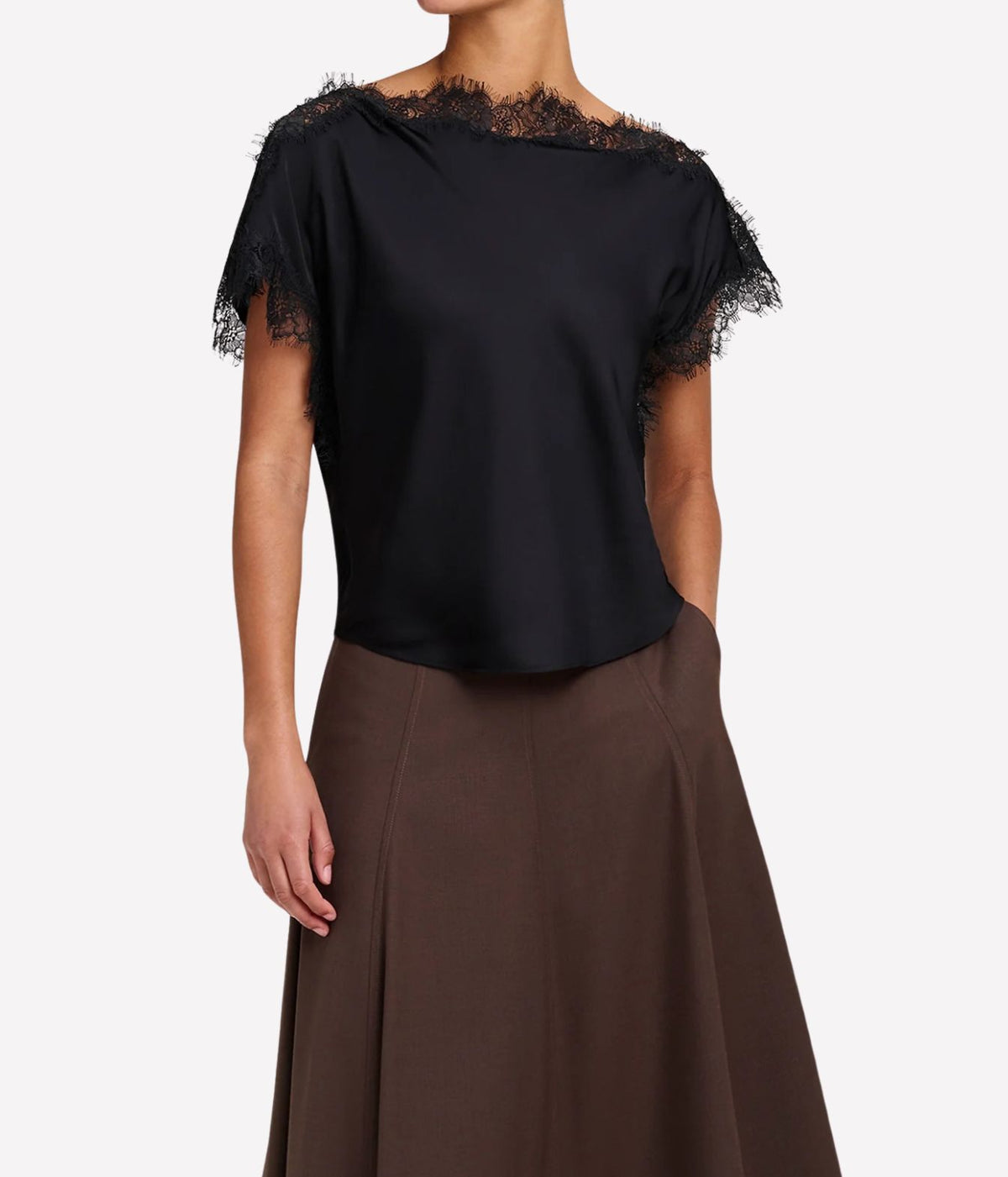 Front view of a model wearing the A.L.C. Luna Top in Black untucked over a chocolate brown pleated midi skirt with one hand in her pocket, showing the eyelash lace neckline and curved hem