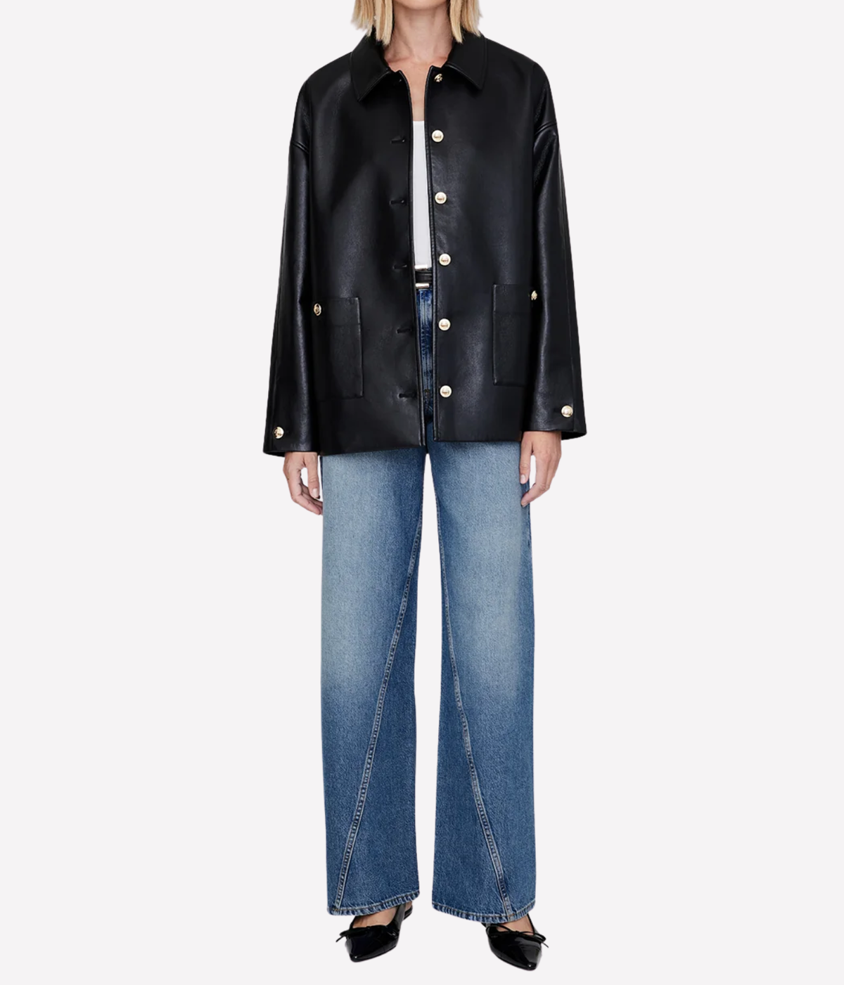 A model wearing a black leather jacket with gold-tone buttons, paired with wide-leg blue jeans and black pointed flats, shot against a plain light gray background.