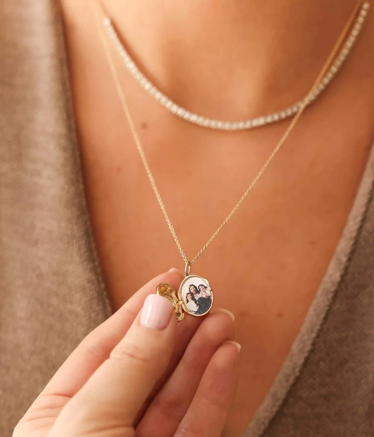 Model holding up the pendant of the Dana Rebecca Designs Teddi Paige Oval Locket Necklace in 14k Yellow Gold, layered with a flat gold chain and a brown knit top
