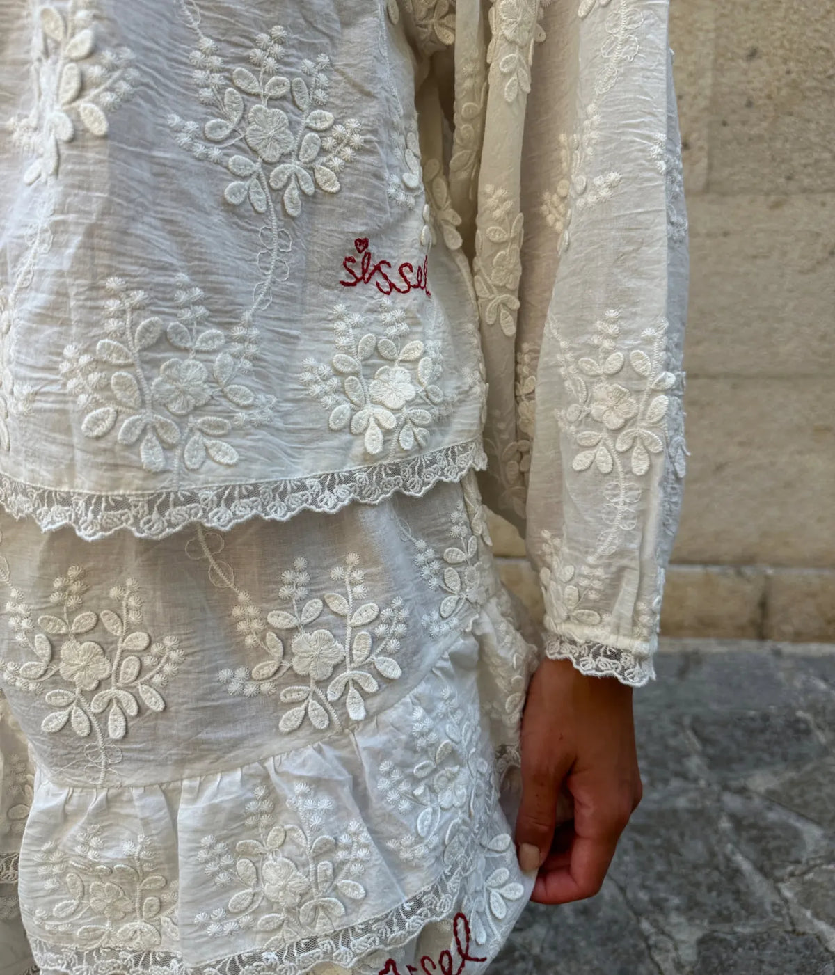 Close-up of the Sissel Edelbo Delphine Top in Off White worn with matching tiered shorts, showing the floral embroidery, lace-trimmed hem and cuff and the red Sissel embroidered signature