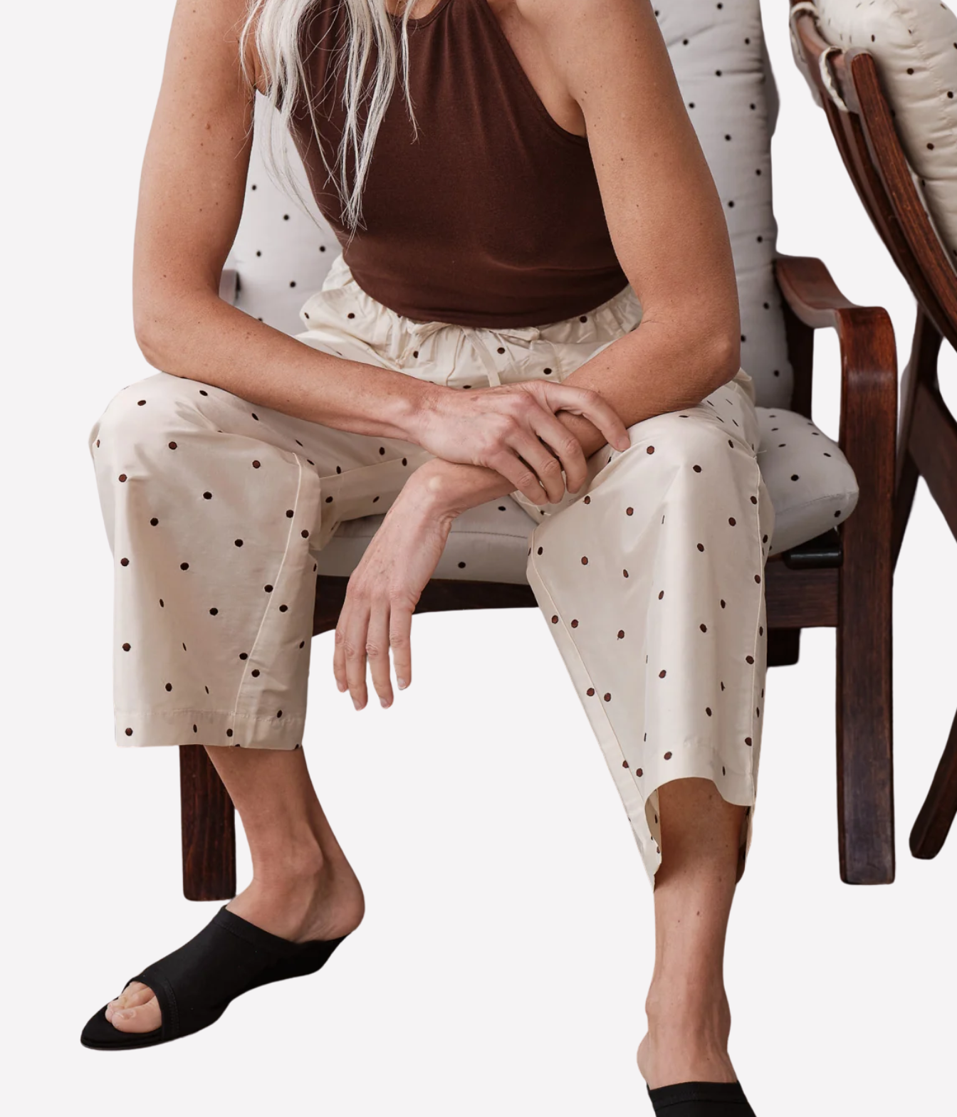 A woman seated on a wooden chair wearing cream wide-leg pants with a dark polka dot pattern, a brown sleeveless top, and black slide sandals against a plain light background.
