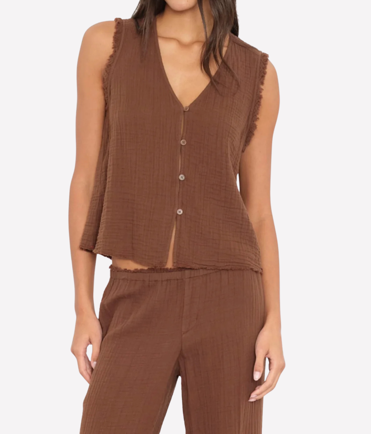 A woman wearing a sleeveless brown textured vest with a button front and frayed edges, paired with matching brown pants, against a light gray studio background.