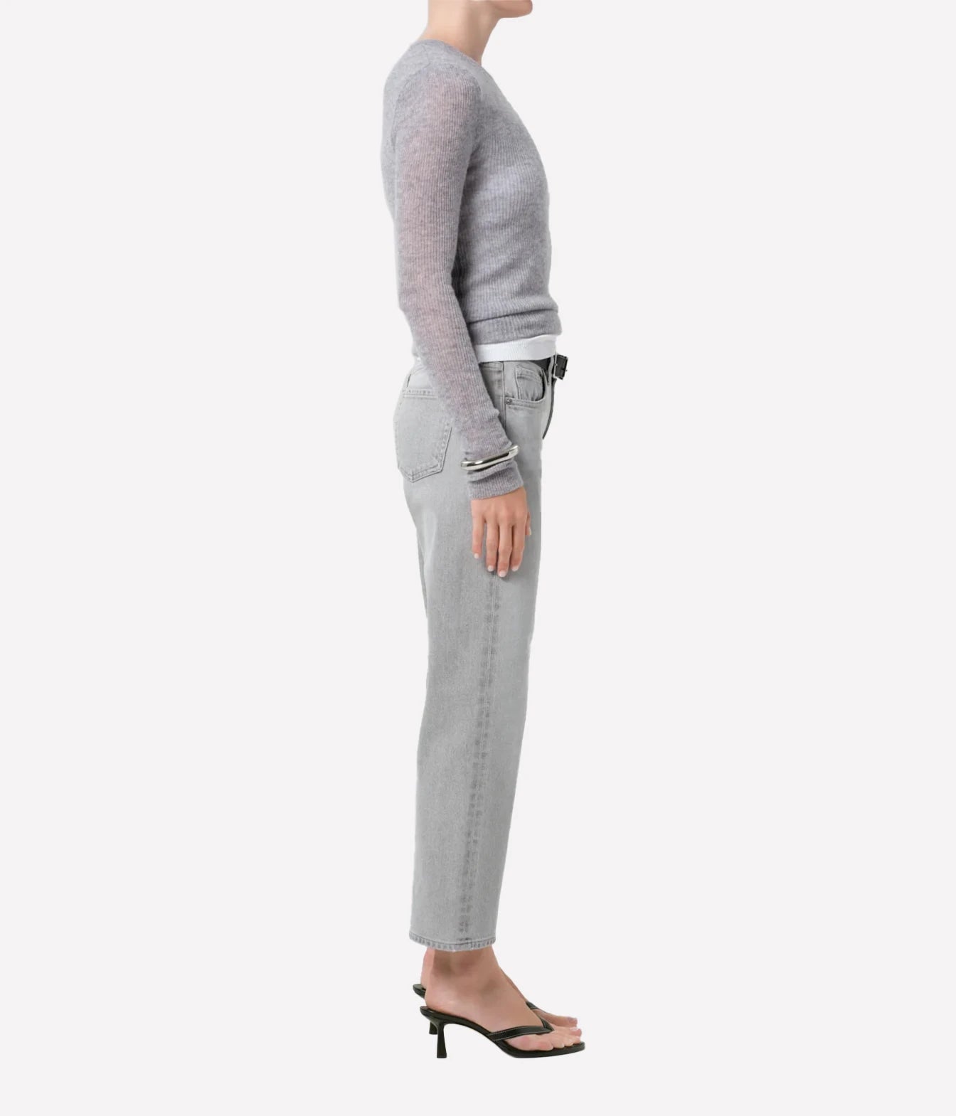 A side-profile view of a model wearing a light gray knit long-sleeve top, light gray slim-fit jeans, and black heeled sandals against a plain light gray background.