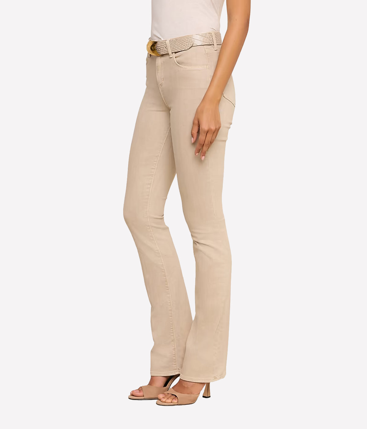 A front side view of a model wearing a pair of the L'AGENCE Selma Bootcut Jean in Biscuit with a matching textured belt, paired with a white top and beige heeled sandals, shown from the waist down.