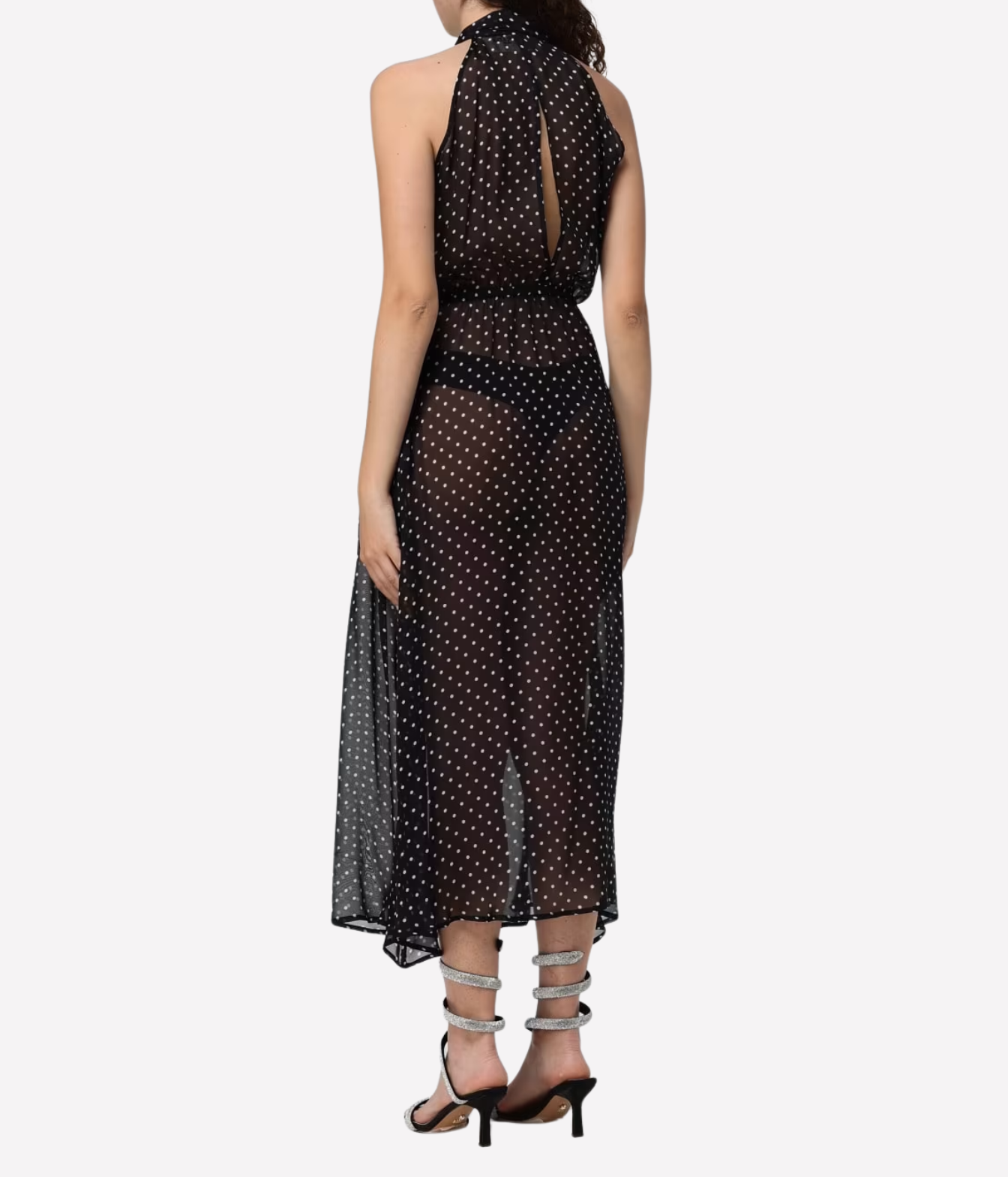 A model from the back wearing a black halter-neck midi dress with white polka dots and a keyhole back design, paired with silver strappy high-heeled sandals against a light gray background.