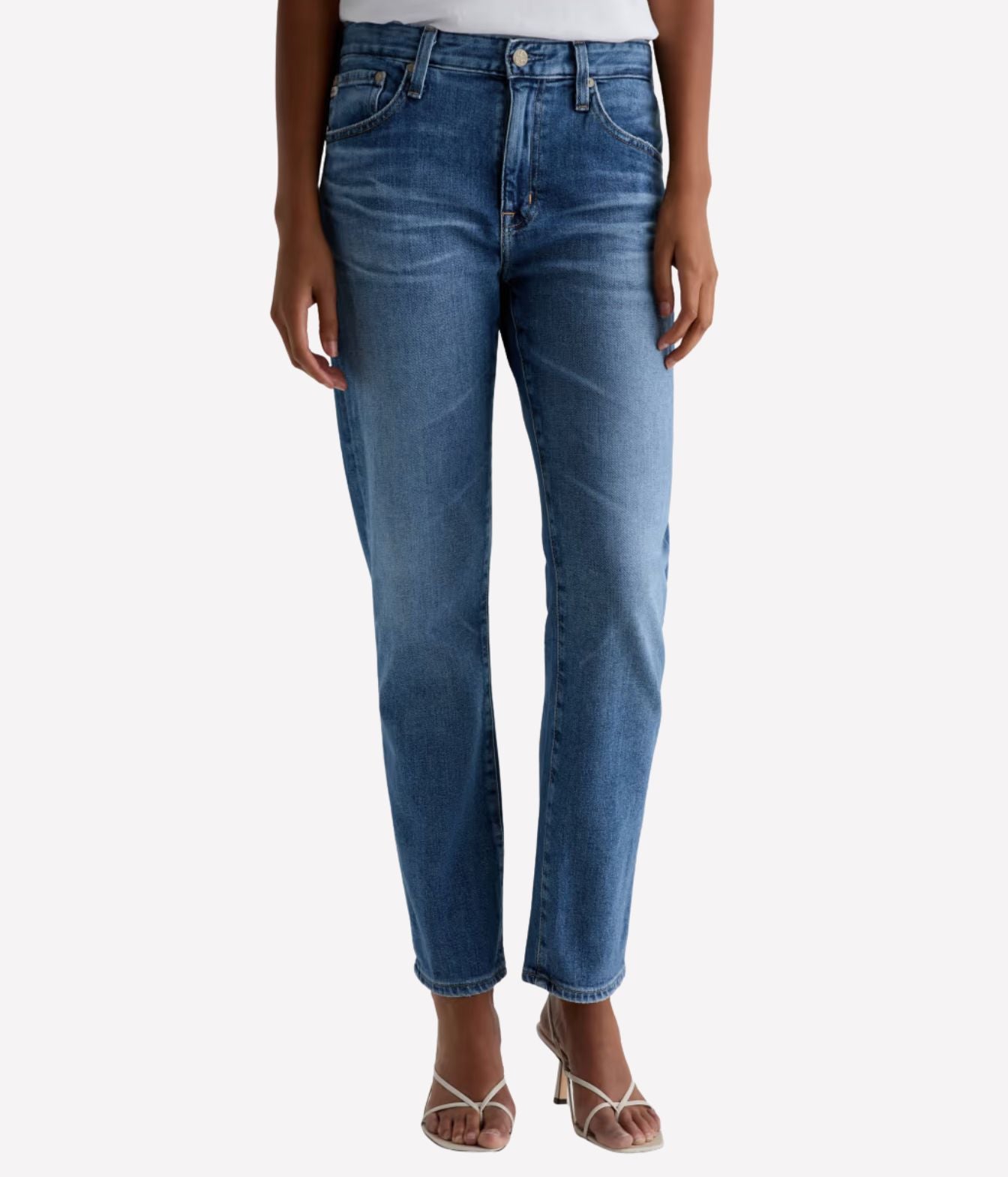 A model wearing a pair of medium wash blue denim jeans with a straight-to-ripped leg, paired with beige strappy heels against a light gray background.
