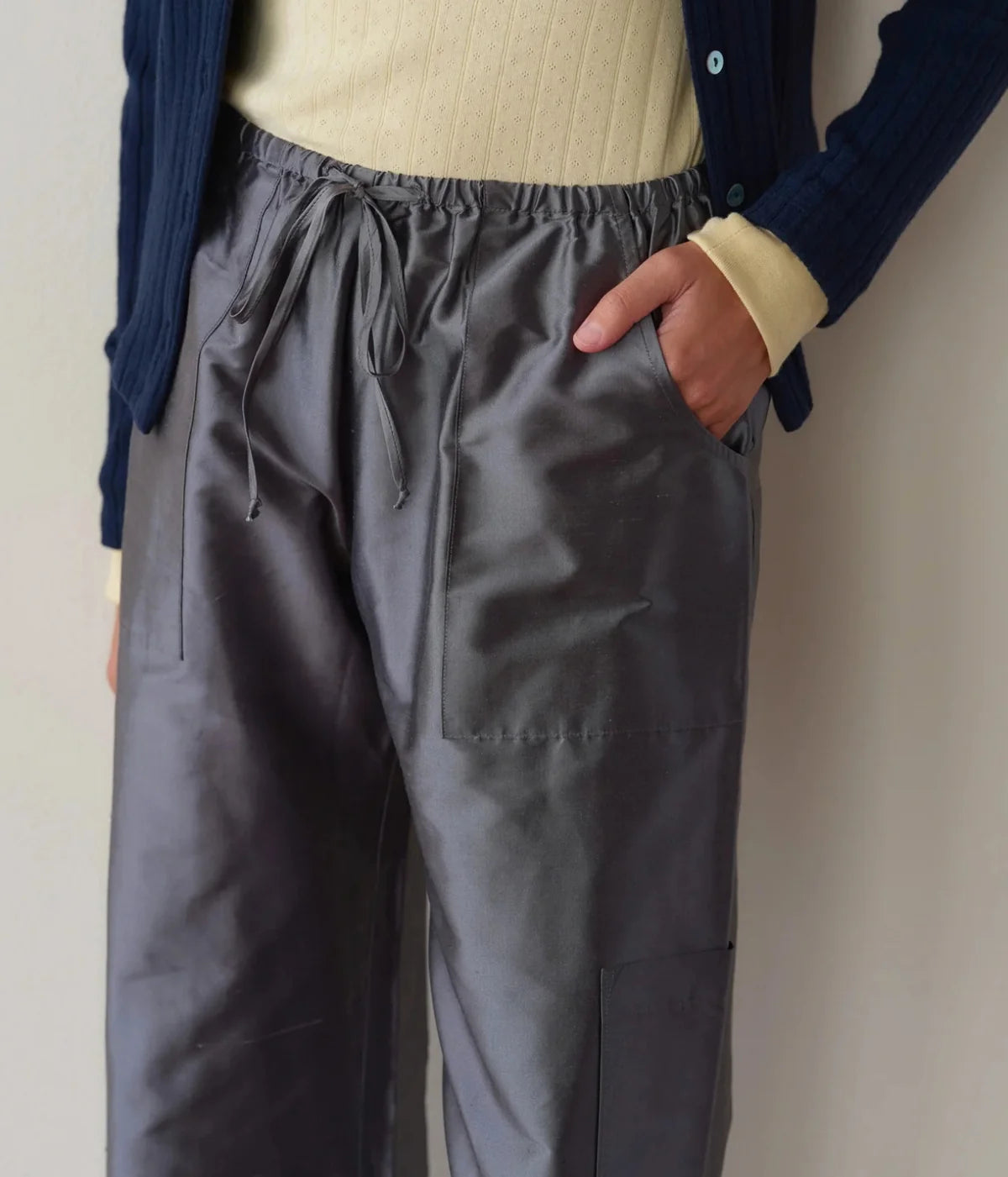 Close-up of a model wearing the DONNI. The Silk Taffeta Cargo Pant in Pewter with a hand in the front pocket, showing the tied drawstring, topstitched patch pockets and the top of the cargo pocket, worn with a pale yellow ribbed top and navy cardigan