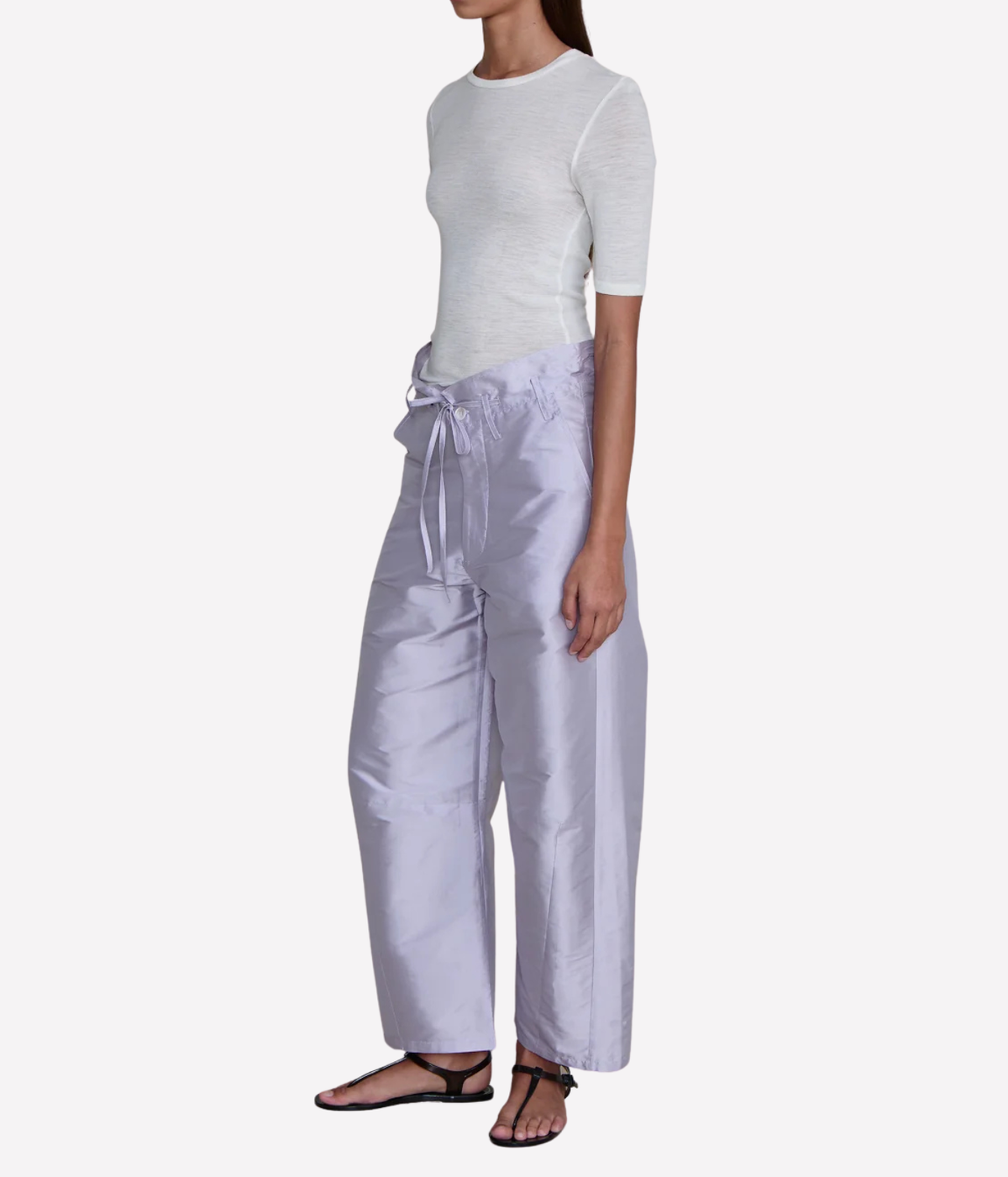 Side profile of a model wearing the Chan Luu Techno Taffeta Barrel Pant in Lilac with a fitted white short-sleeve top and black thong sandals, showing the curved barrel-leg shape and cropped length