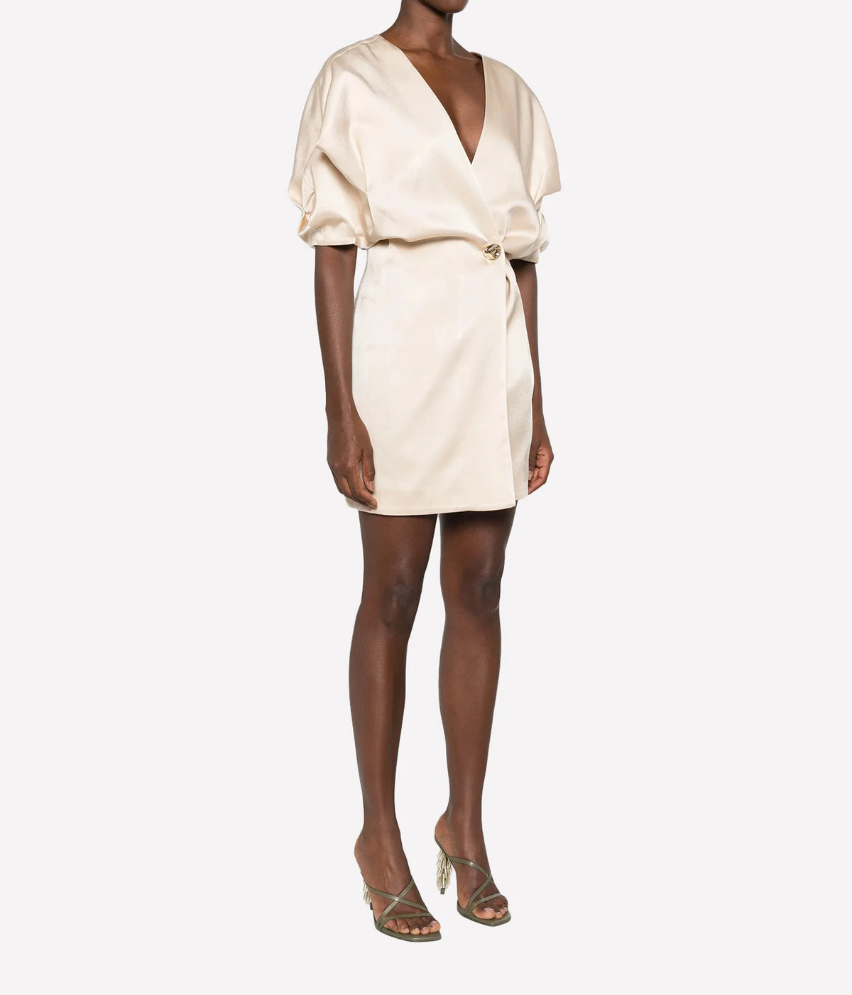 Three-quarter view of a model wearing the Raquel Diniz Anastasia Short Dress in Vanilla with strappy heeled sandals, showing the draped sleeves and wrap front