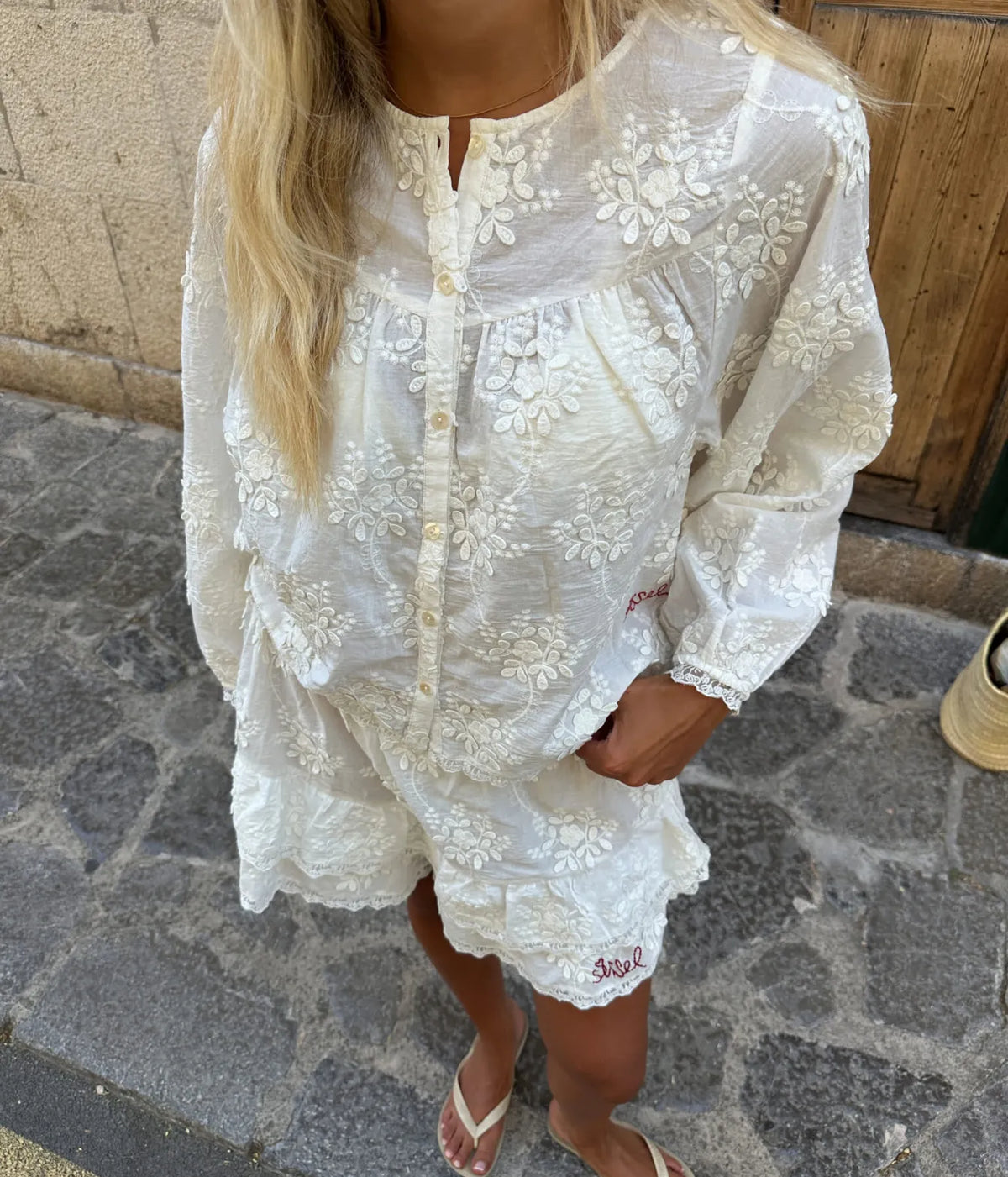Model photographed from above wearing the Sissel Edelbo Delphine Top in Off White buttoned over matching ruffle shorts with tan flat sandals, showing the embroidered floral panels, gathered yoke and lace-trimmed cuffs
