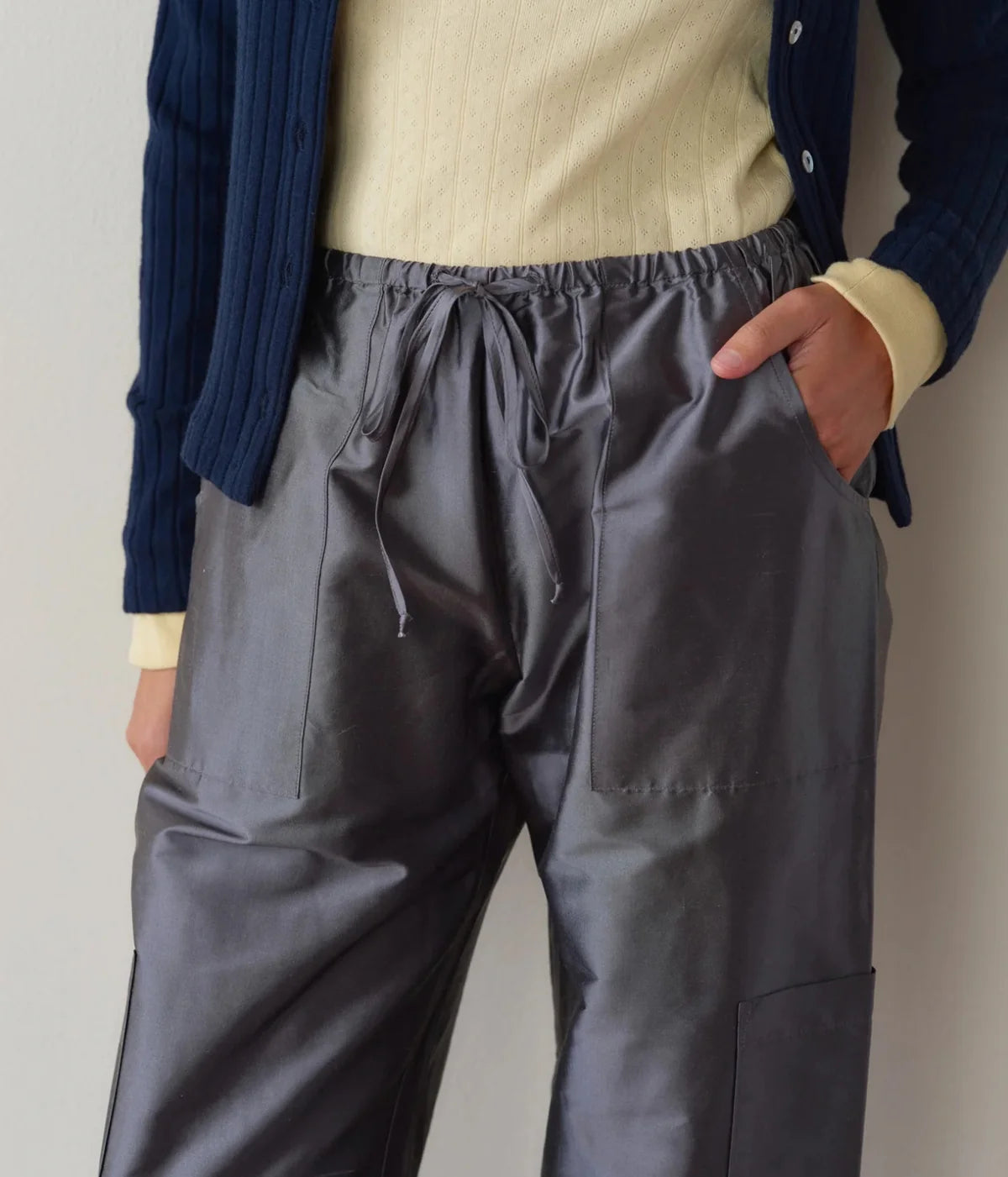 Detail of the DONNI. The Silk Taffeta Cargo Pant in Pewter, showing the sheen of the silk taffeta, the gathered drawstring waist and stitched front pockets, worn with a pale yellow pointelle top and navy cardigan