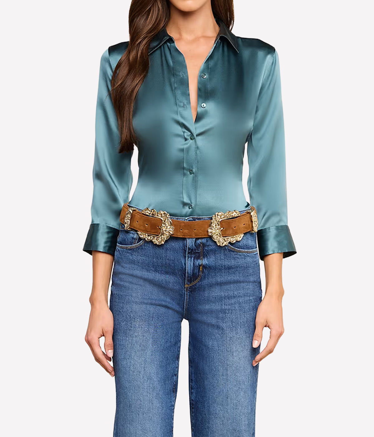 A woman wearing a cadet blue silk button-down shirt with rolled cuffs, tucked into blue jeans and styled with a brown leather belt featuring large ornate gold buckles.