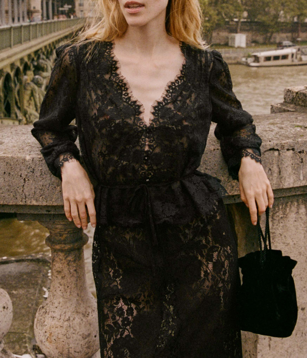 Front view of a model wearing the DÔEN Amaline Top In Black with the waist tie knotted over a matching black lace midi skirt, holding a black velvet drawstring pouch bag