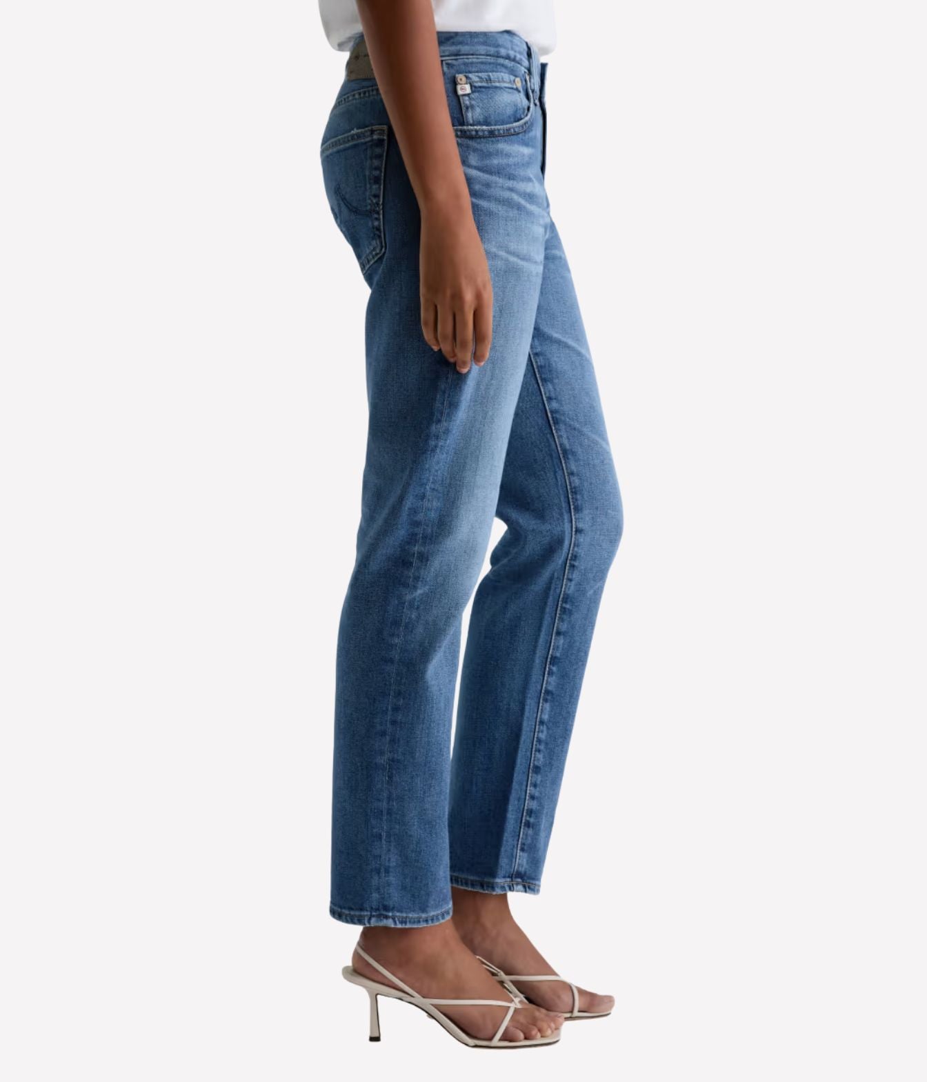 A side-profile view of a person wearing medium-wash blue denim jeans with a straight-to-regular leg fit, paired with clear-strap high-heeled sandals against a light gray studio background.