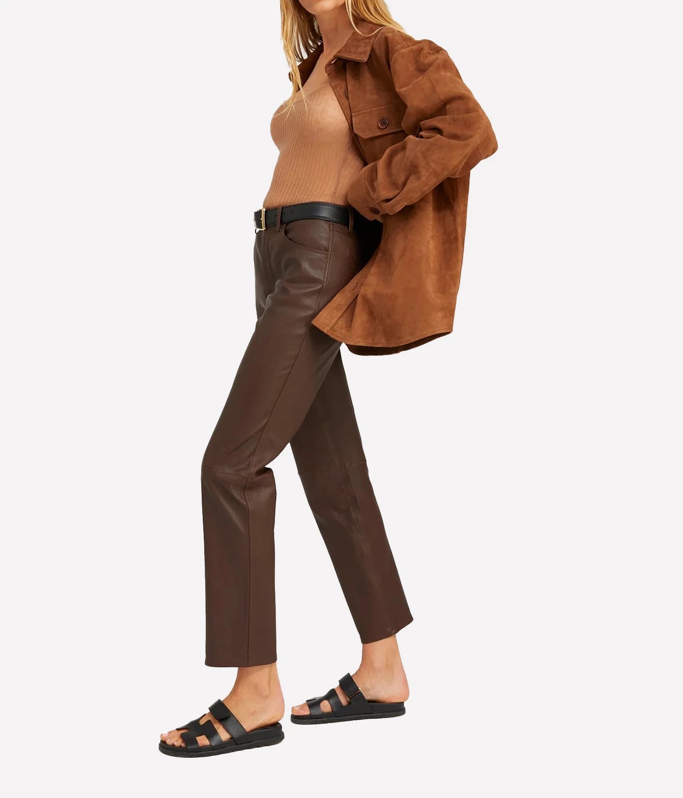 A model wearing brown faux-leather cropped trousers, a tan ribbed knit top, a brown suede overshirt, and black slide sandals, shown from the waist down against a light gray background.
