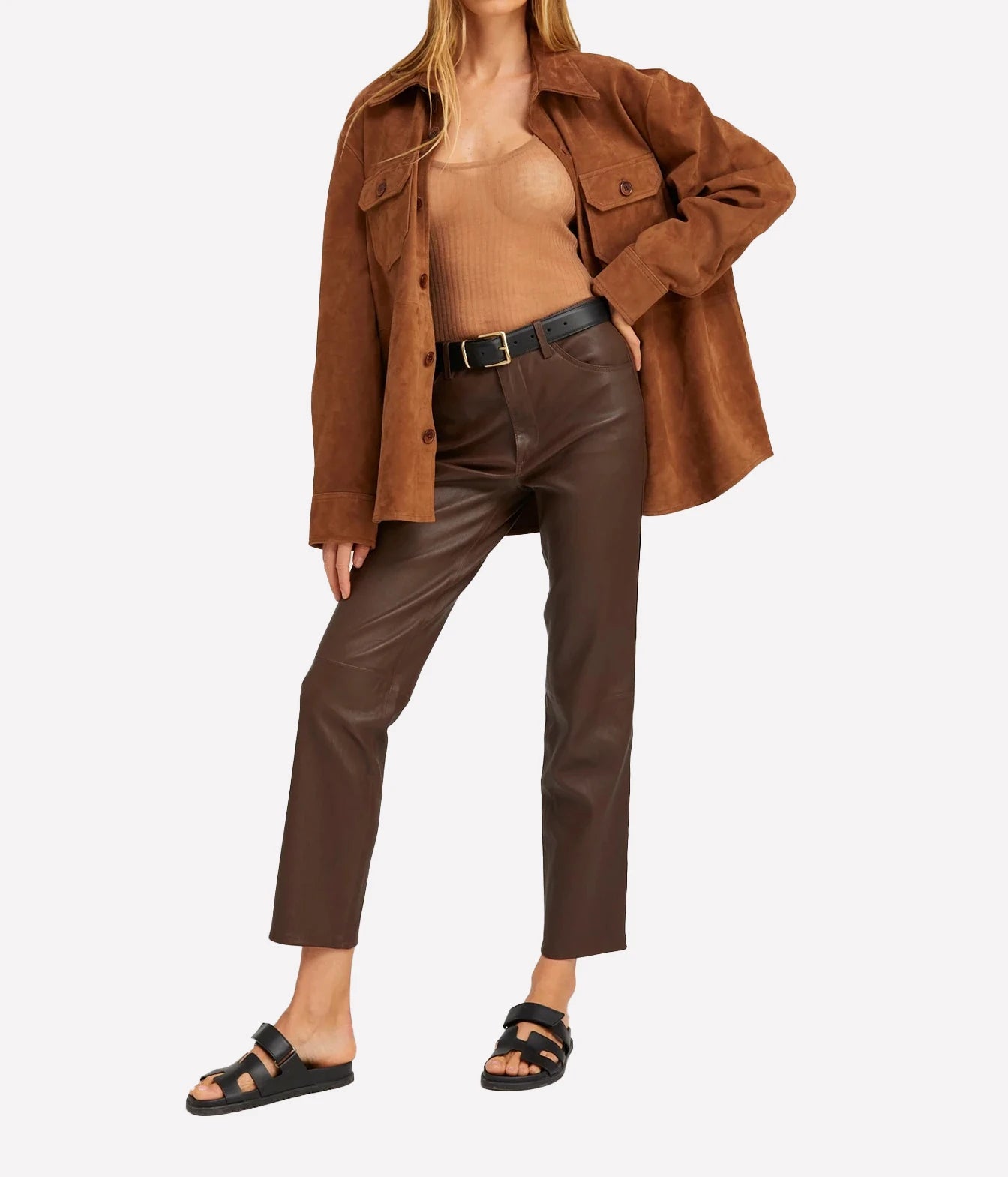 A model wearing brown cropped faux-leather pants with a black belt, styled with a tan ribbed tank top, an oversized brown suede jacket, and black slide sandals, set against a plain light gray background.