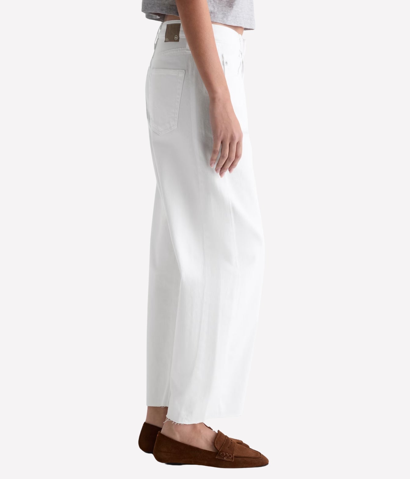 A side profile view of a person wearing white wide-leg cropped jeans with brown loafers, shown against a plain light gray studio background.