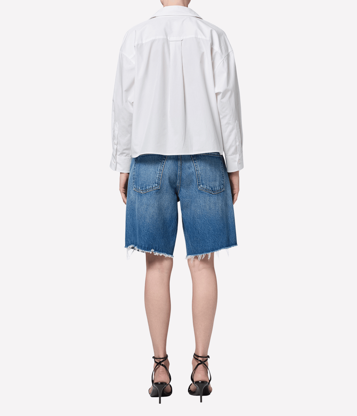 Back view of a model wearing the Citizens of Humanity Ayla Short in Chaya with a loose white shirt and black strappy heeled sandals, showing the back pockets and frayed raw hems