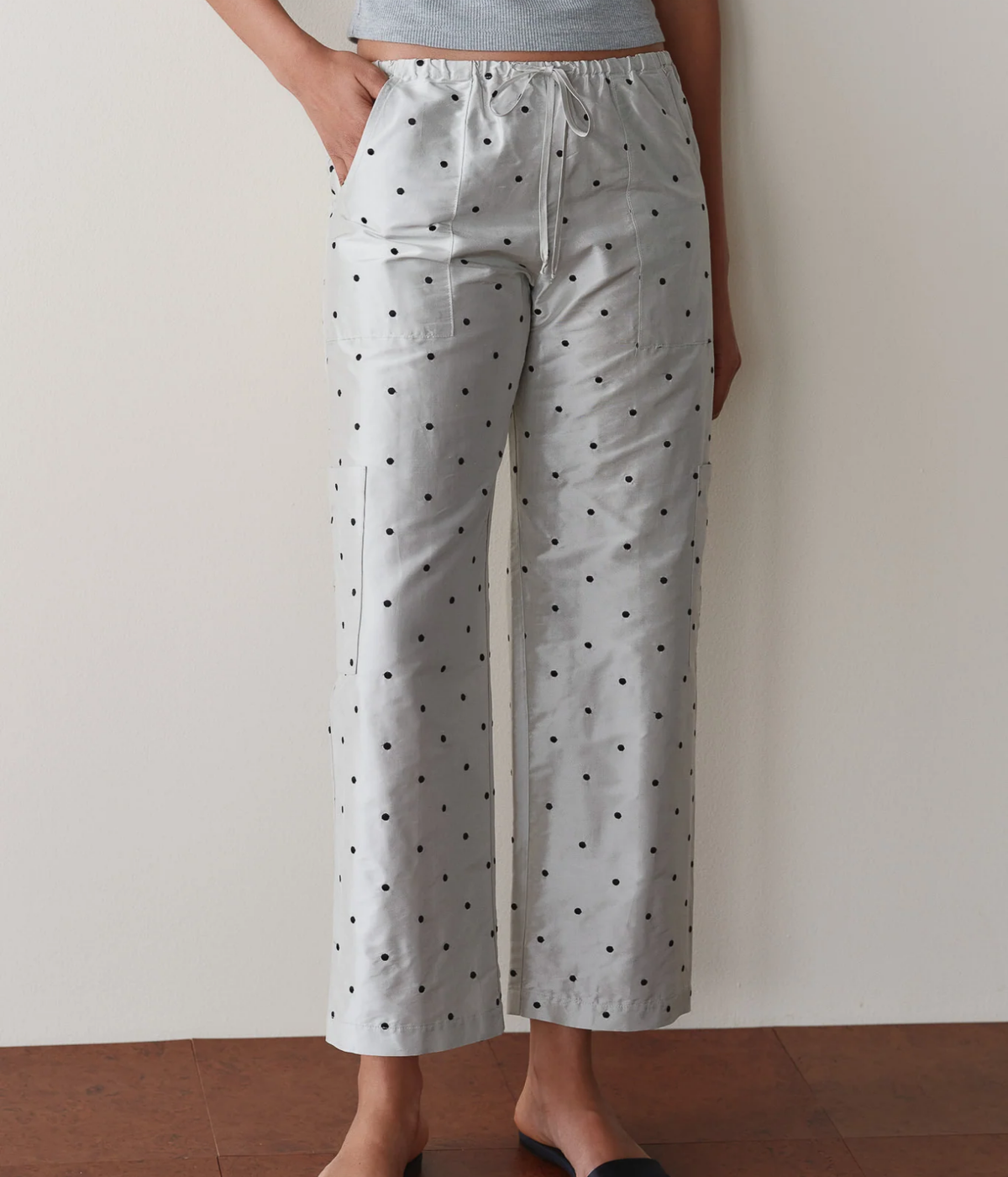Model wearing the DONNI. The Silk Taffeta Cargo Pant in Silver Polka Dot with a hand in the front pocket, styled with a grey tank top and black slide sandals