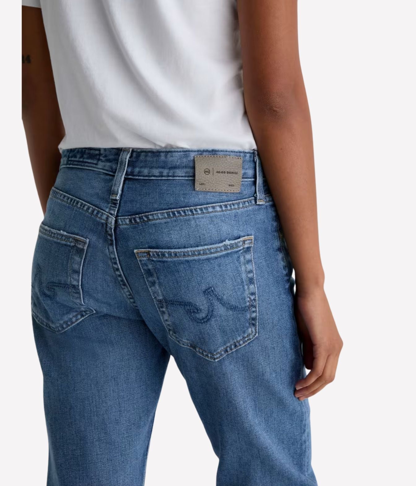 Rear view of a person wearing blue denim jeans with a white top, showing the back pockets and waistband label against a light gray background.