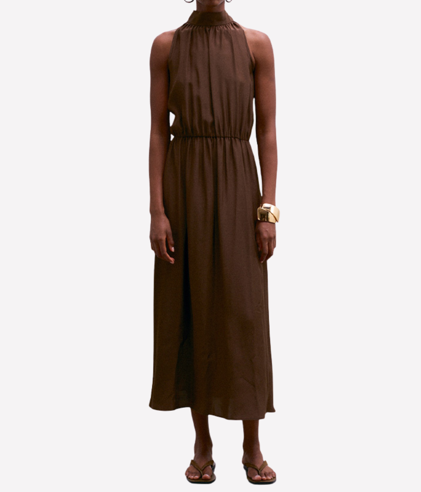 A model wearing a sleeveless, high-neck, midi-length brown dress with a gathered waist, paired with brown sandals and a gold bracelet, set against a plain light gray background.