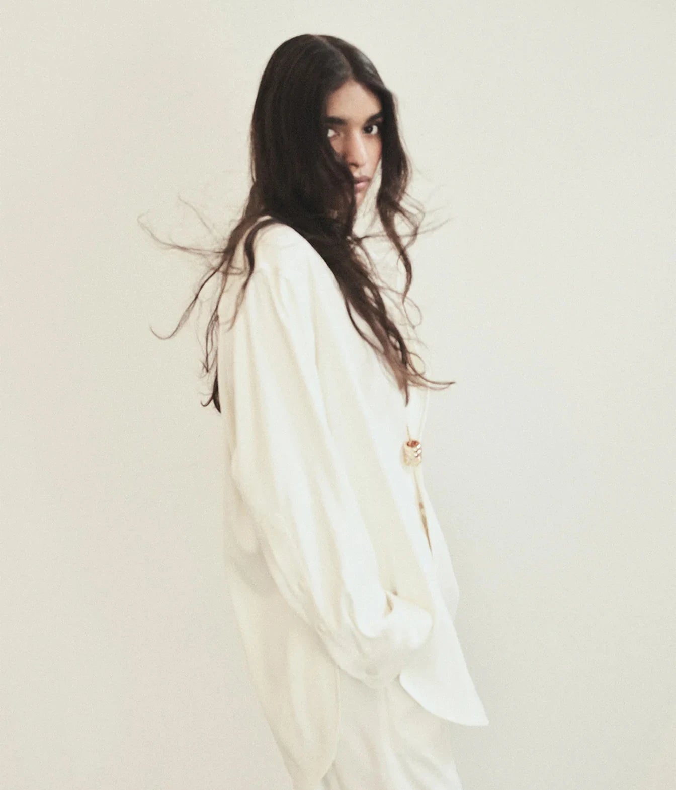 A woman with long, dark wavy hair wearing an oversized white long-sleeve top and matching white bottoms, photographed against a plain off-white studio background.