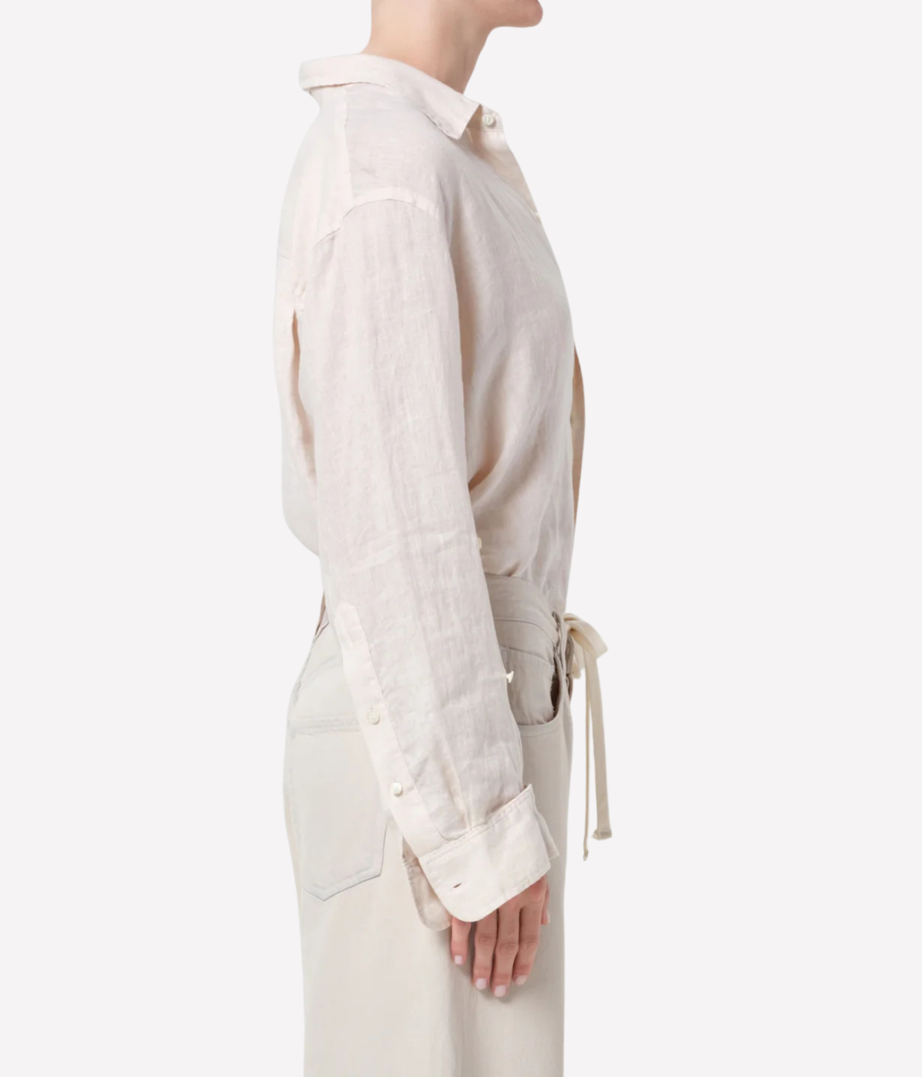A side-profile view of a model wearing a light beige linen shirt with a collar, rolled cuffs, and a matching waist tie, paired with light beige trousers against a plain white background.