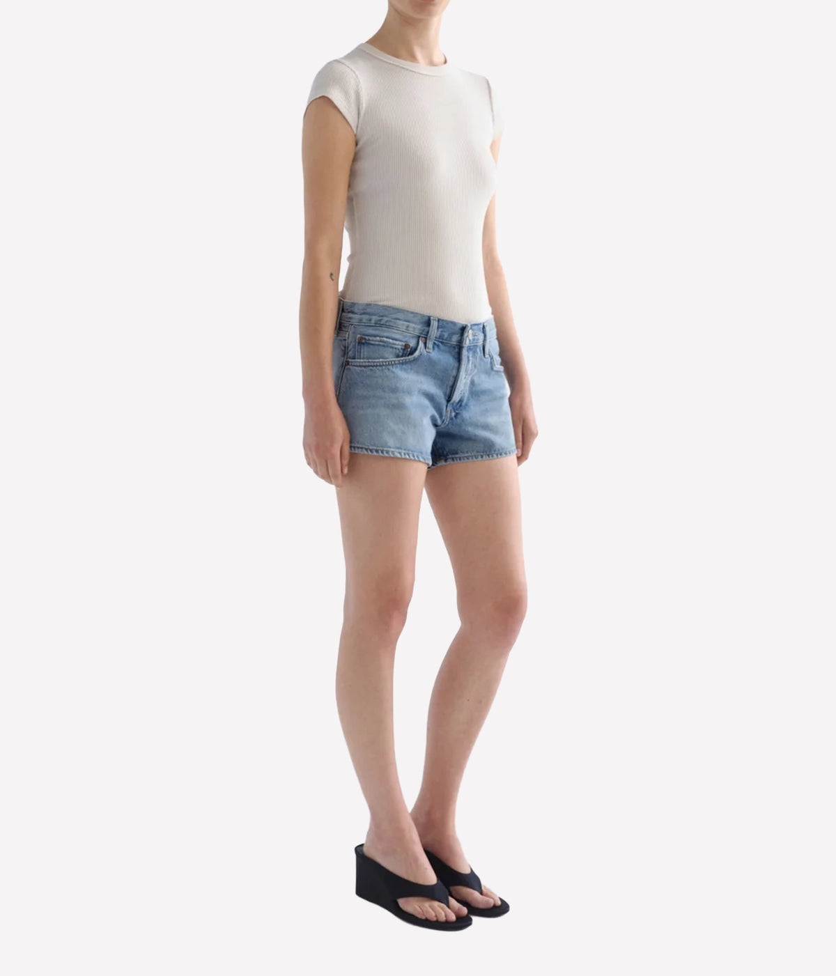A model wearing a cream ribbed short-sleeve top, light-wash blue denim shorts, and black wedge sandals, Agolde Low Rise Parker Short in Replica