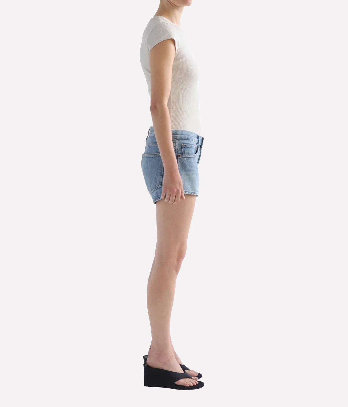 A side-profile view of a model wearing light-wash denim shorts, Agolde Low Rise Parker Short in Replica, a white short-sleeve top, and black wedge sandals