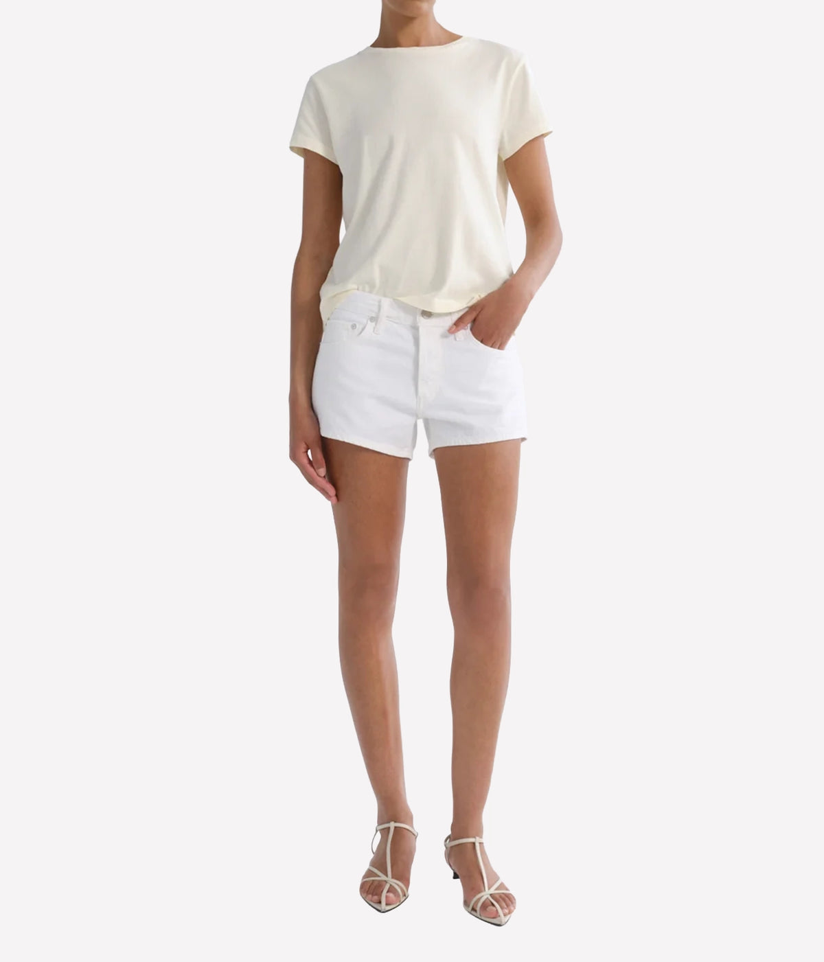 Model wearing the Agolde Low Rise Parker Short in Marshmallow with a cream short-sleeve tee, one hand in the front pocket, and nude strappy heeled sandals