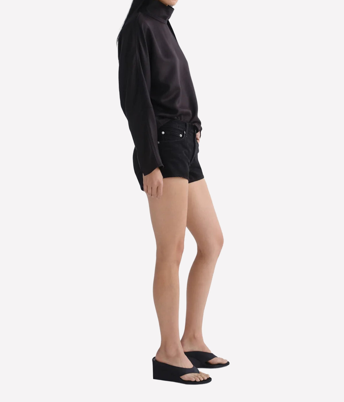 A model shown from the side wearing a black long-sleeve mock-neck top, black denim shorts, and black wedge slide sandals - Agolde Low Rise Parker Shorts in Phase