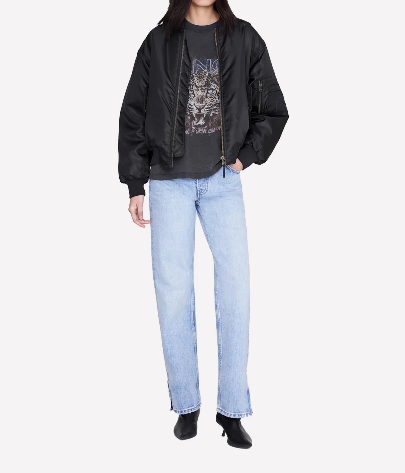 A model wearing a black bomber jacket over a black graphic t-shirt, paired with light-wash blue straight-leg jeans and black ankle boots, shot against a light gray studio background.