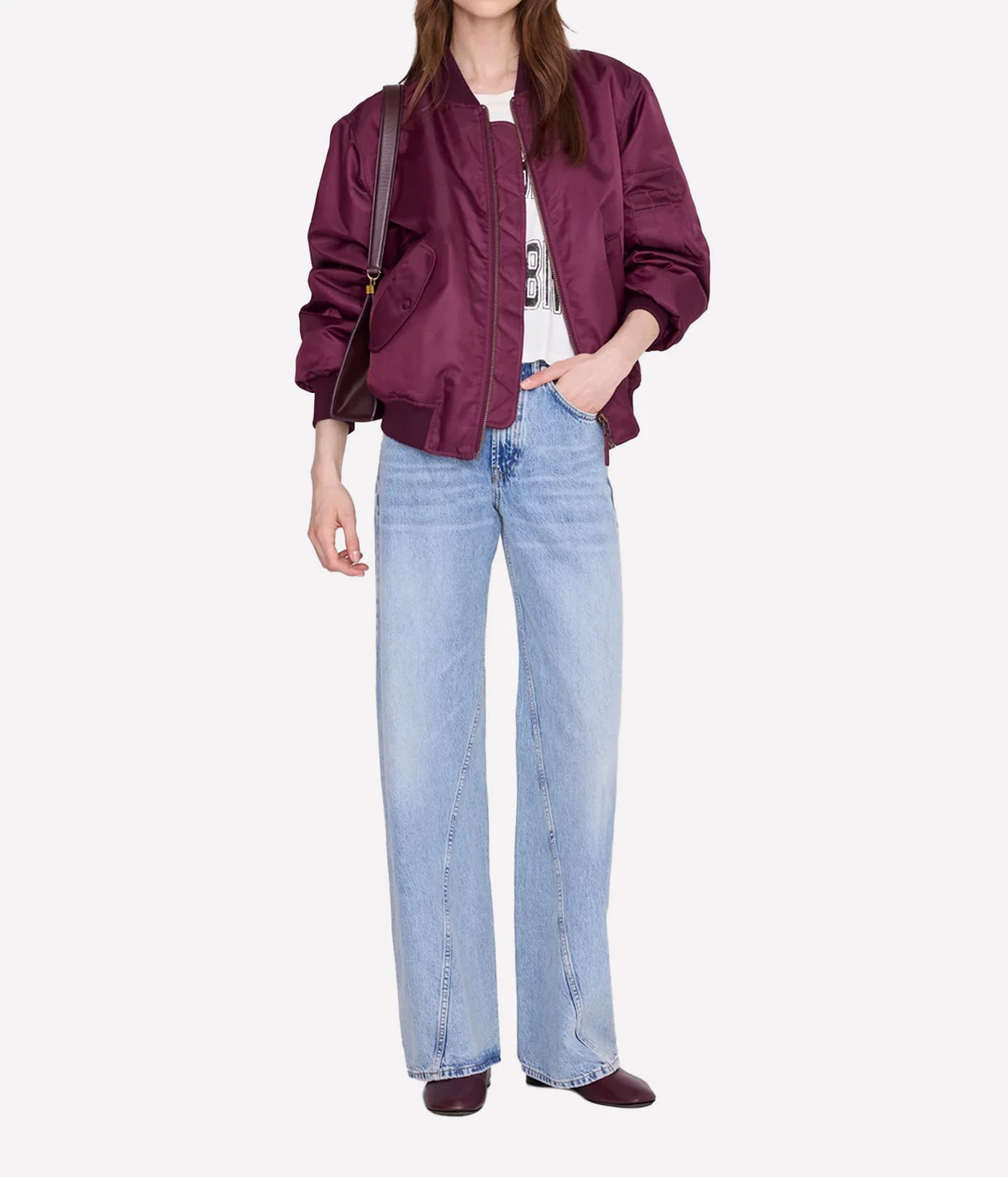 A model wearing a shiny burgundy bomber jacket, light-wash wide-leg blue jeans, a white top, and burgundy shoes, standing against a light gray background.
