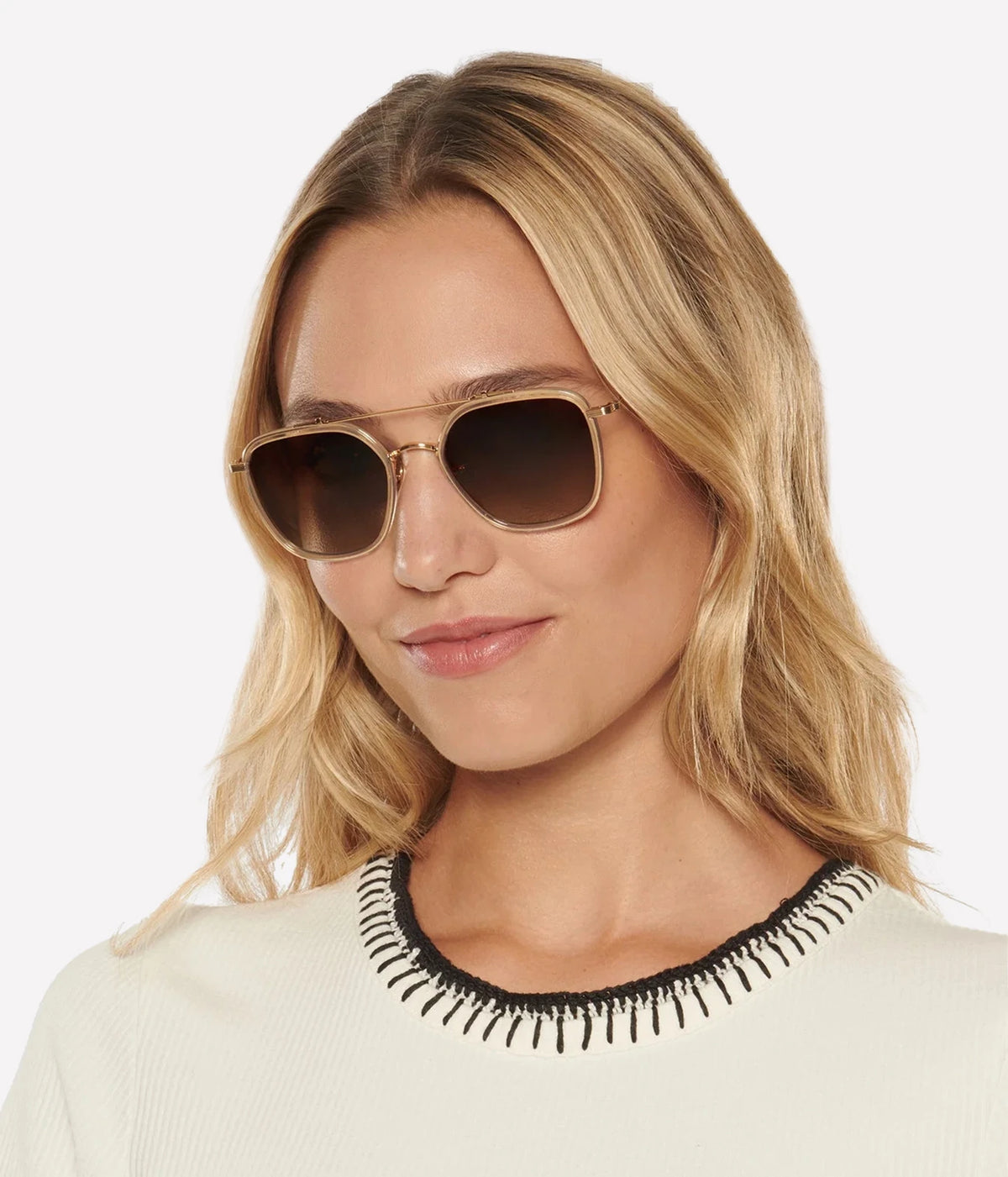 Model with wavy blonde hair wearing the KREWE Austin Sunglasses in 18K Titanium Champagne with a cream knit trimmed in black and white