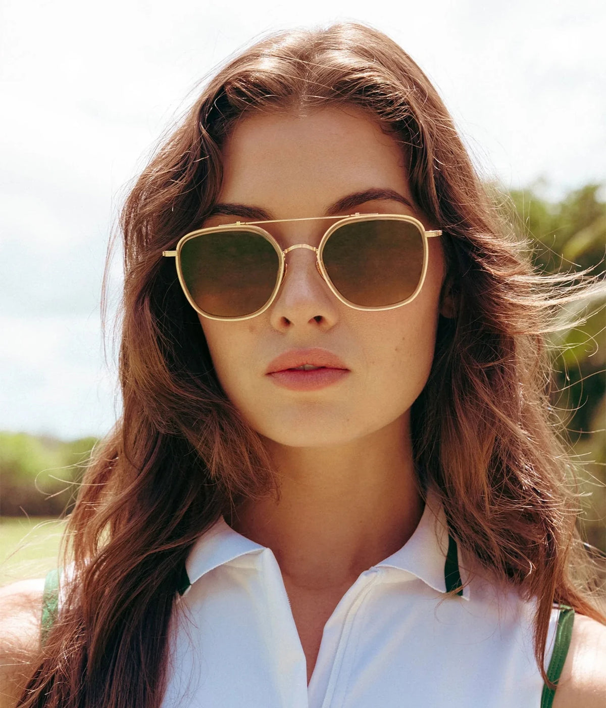 Model with long brown hair wearing the KREWE Austin Sunglasses in 18K Titanium Champagne with a white top, front view