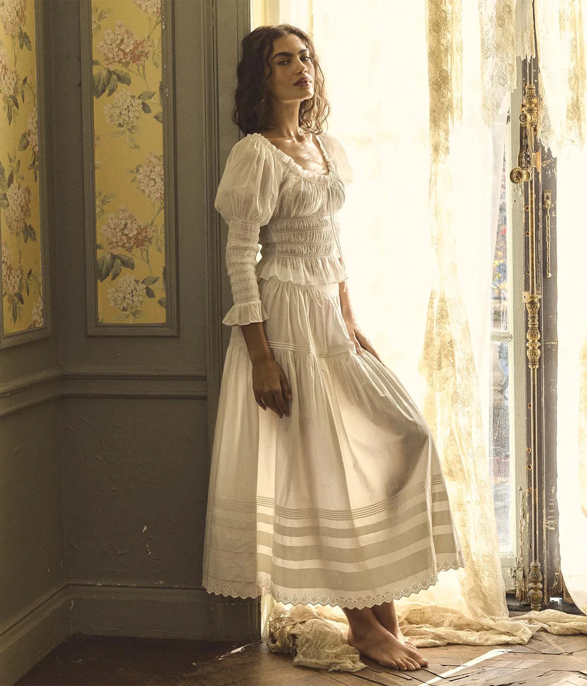 A woman stands barefoot in a sunlit room wearing a long white puff-sleeve top paired back with a maxi cotton skirt with horizontal stripes near the hem. She is posed beside a window with sheer curtains, with floral wallpaper visible on the wall.