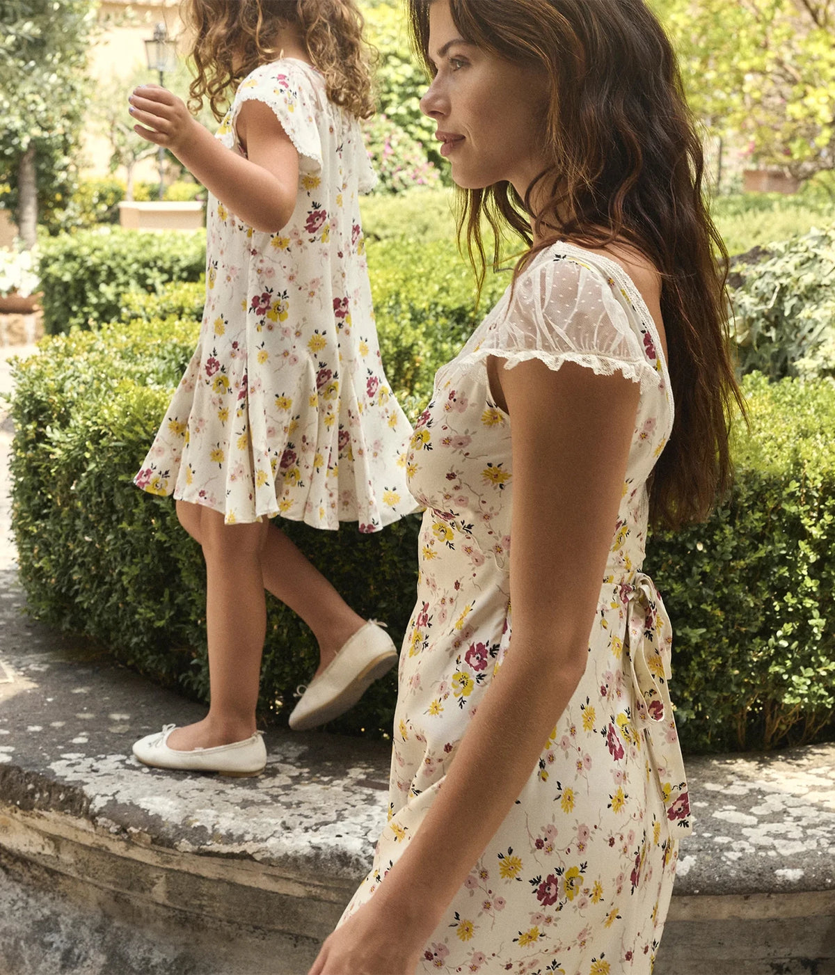 Side profile of a model wearing the DÔEN Corelia Dress In Sweetbriar Floral turning towards a young girl in a matching dress, showing the cap sleeve and fluid skirt