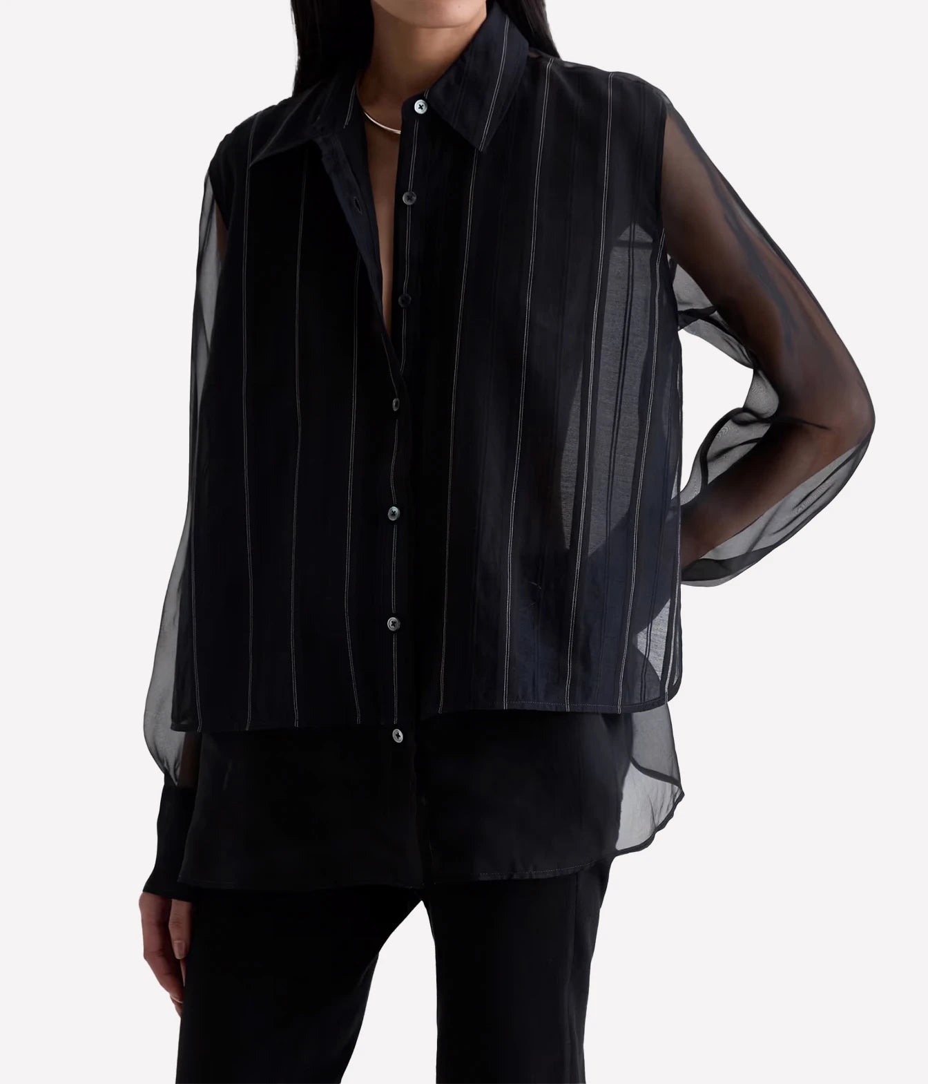 A model wearing a black sheer long-sleeve button-up shirt with vertical white pinstripes and a cropped sleeveless overlay, paired with black pants against a light gray background.