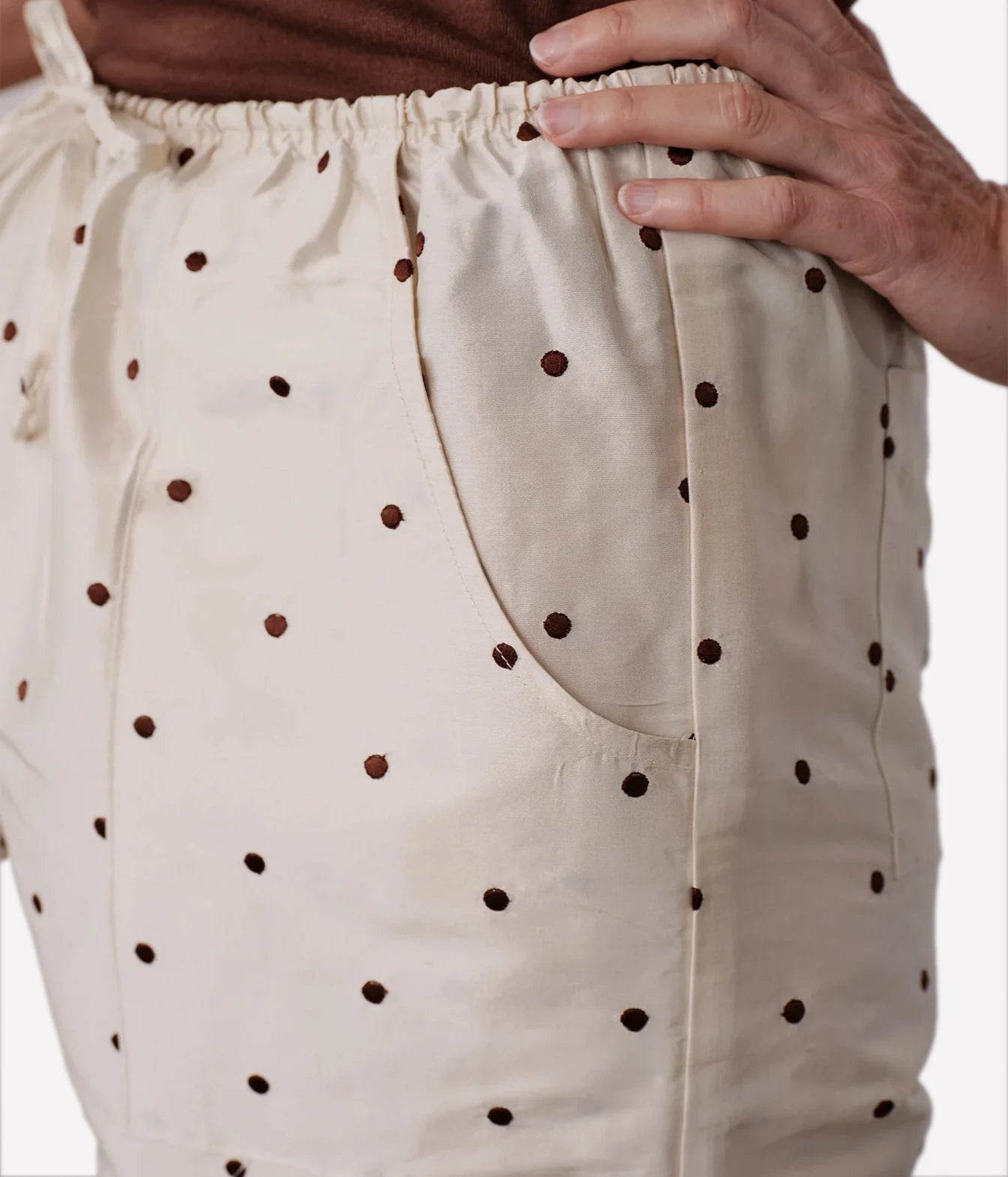 Close-up of a person wearing cream-colored drawstring shorts with dark brown polka dots, showing the fabric texture, side pocket, and hand adjusting the waistband.