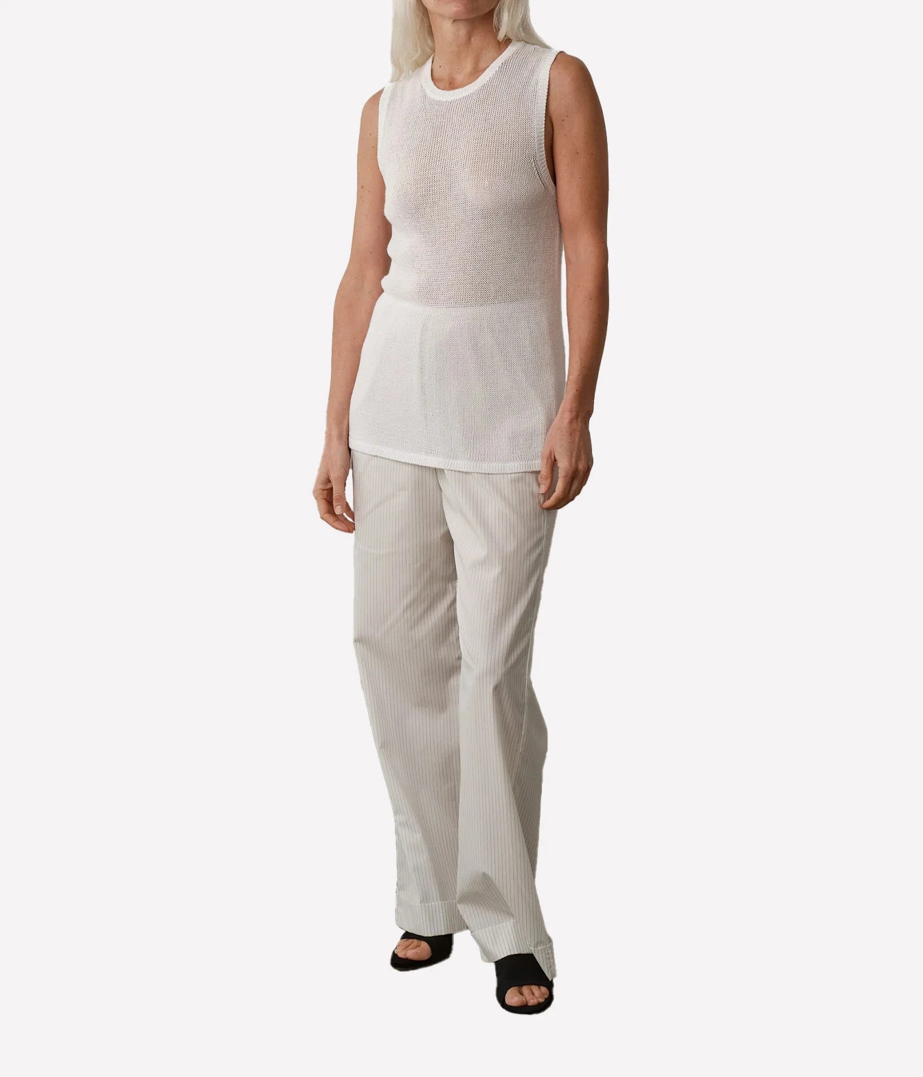 A model wearing a white sleeveless knit top and light beige wide-leg trousers, paired with black slide sandals, against a plain light gray background.