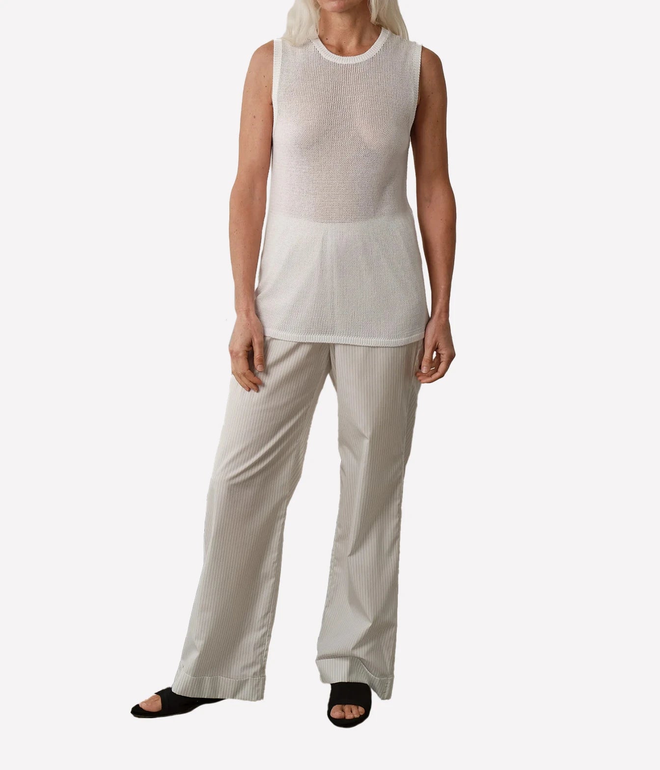 A model wearing a sleeveless white knit top and light beige wide-leg trousers, paired with black slide sandals, against a plain light gray background.