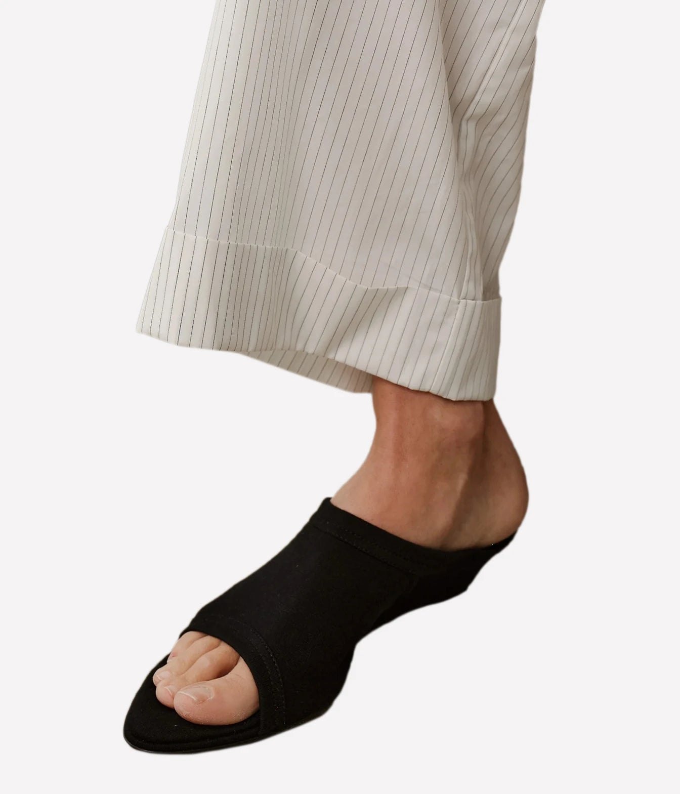 A close-up of a person's foot wearing a black open-toe slip-on sandal, paired with off-white pinstriped trousers, against a plain light gray background.
