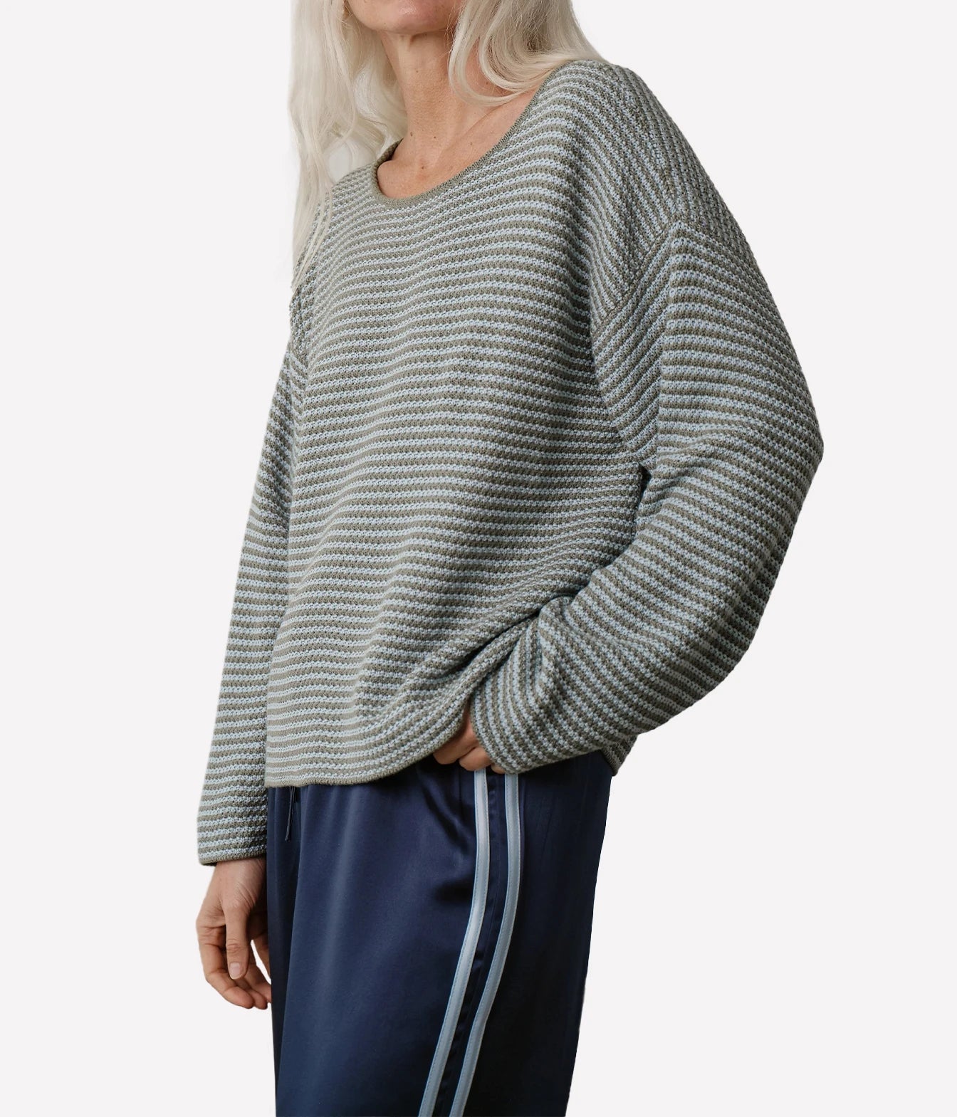 A woman wearing a textured, long-sleeve knit sweater with a gray and white houndstooth-like pattern, paired with navy blue satin track pants with white side stripes, against a plain light gray background.
