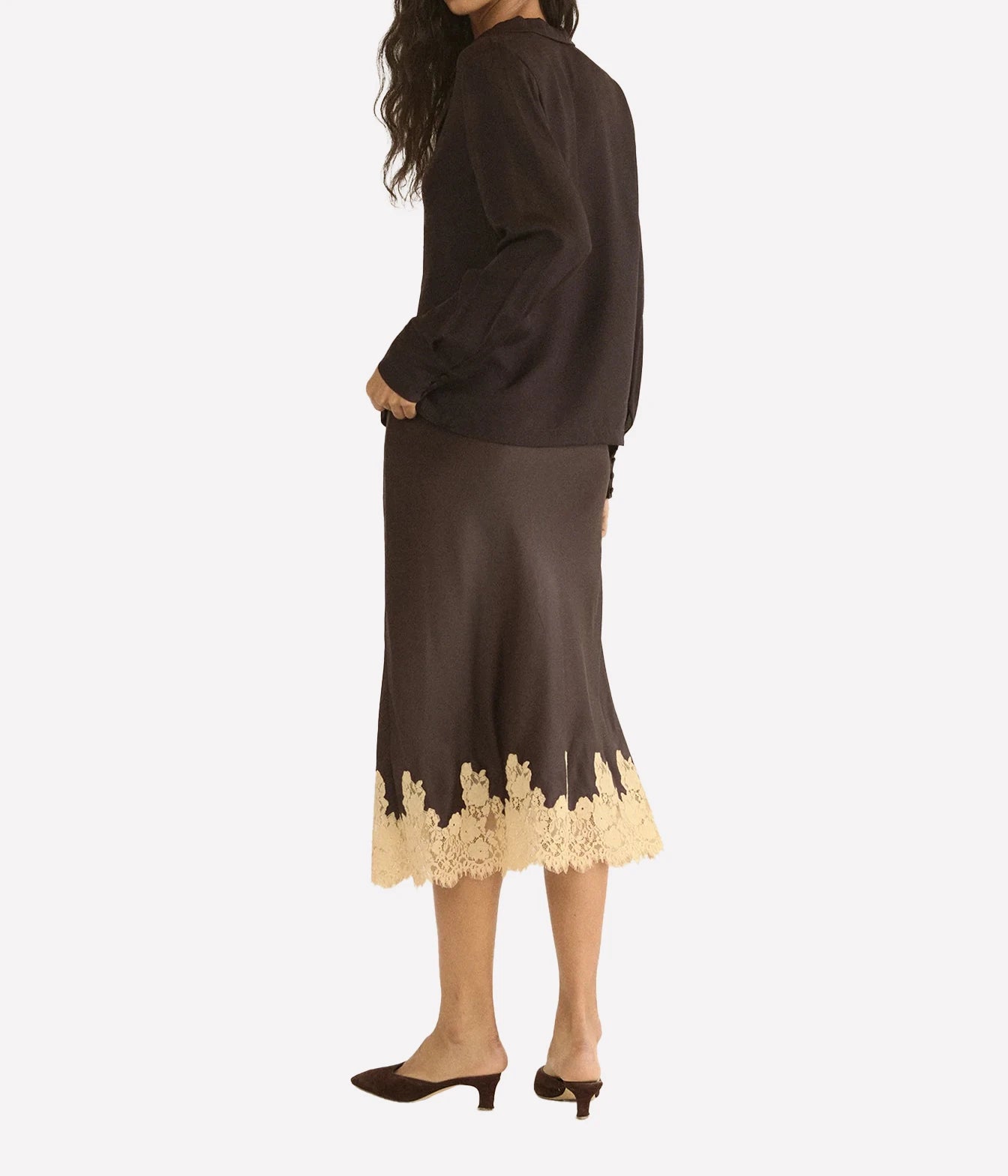 A woman shown from the back wearing a dark brown long-sleeve blouse and matching midi skirt with a beige floral lace hem, paired with dark brown mule heels, against a light gray studio background.